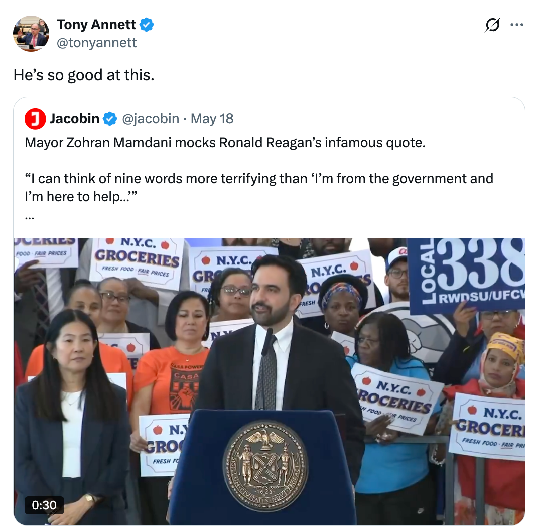 Man in a suit speaks at a podium surrounded by people holding signs about NYC groceries, mocking a Ronald Reagan quote