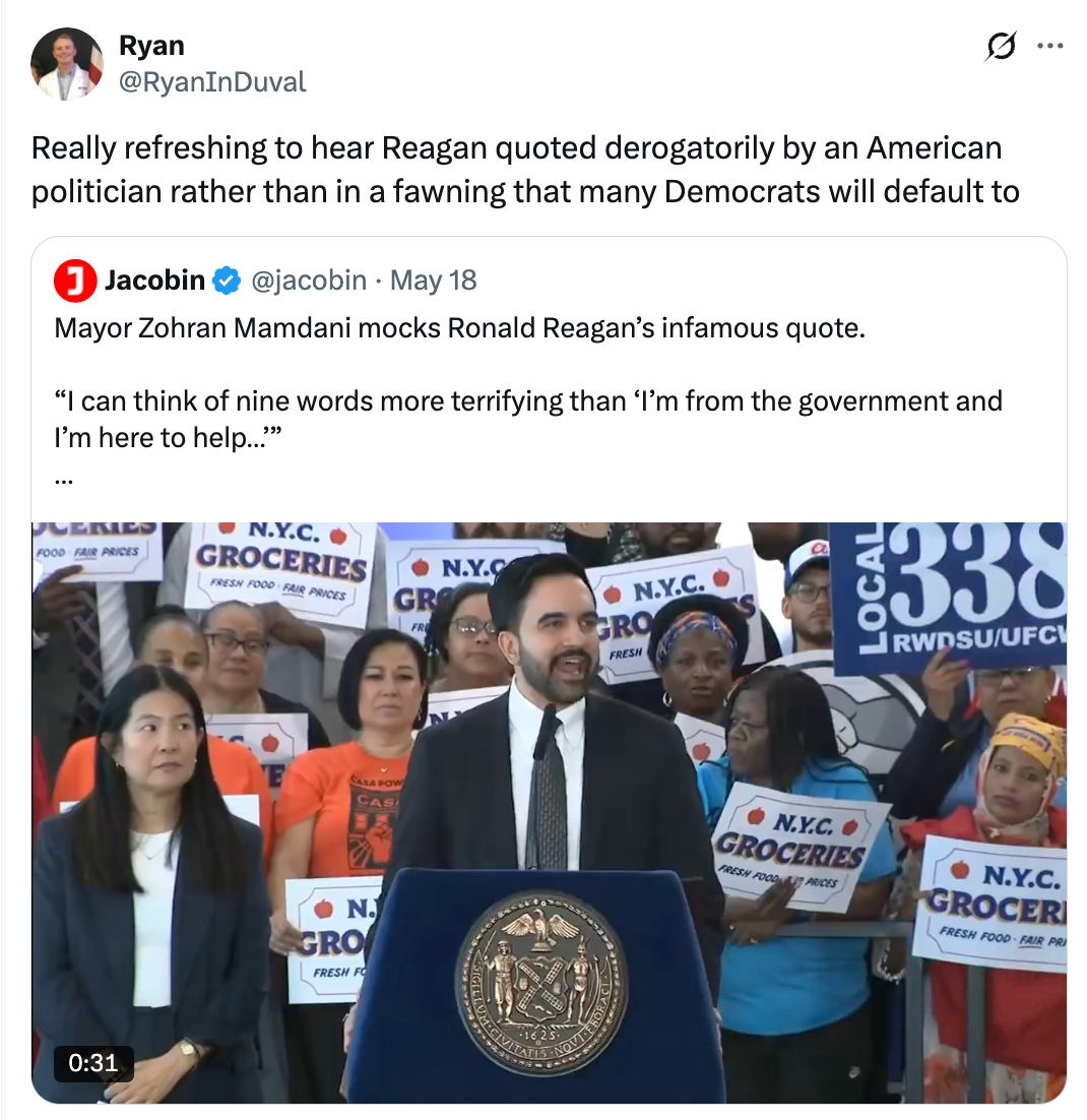 Person at podium mocking Reagan quote; supporters in background hold signs about NYC groceries and pricing