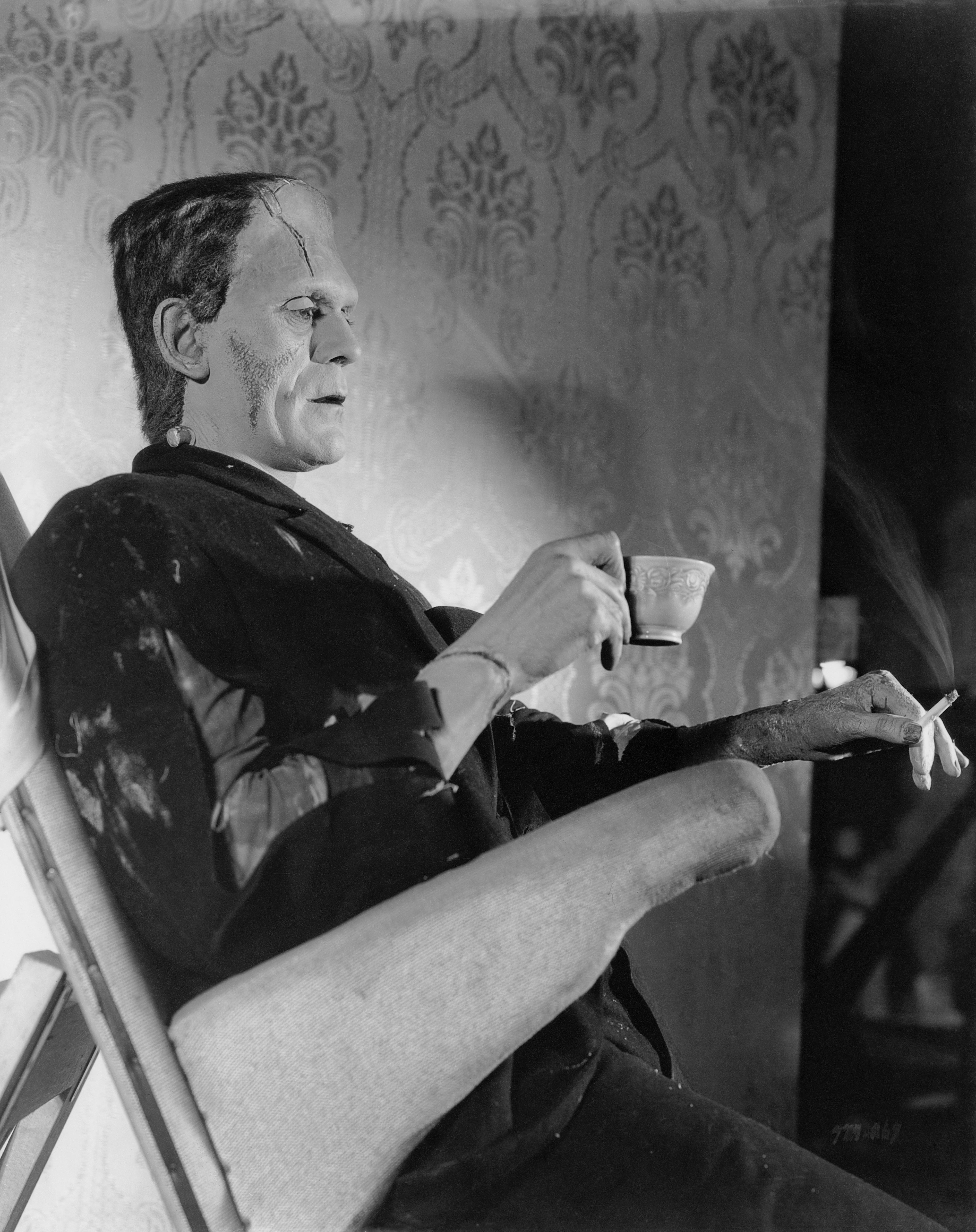 Boris Karloff in Frankenstein makeup relaxing on set, holding a teacup and cigarette, seated in a lounge chair with a textured wall backdrop