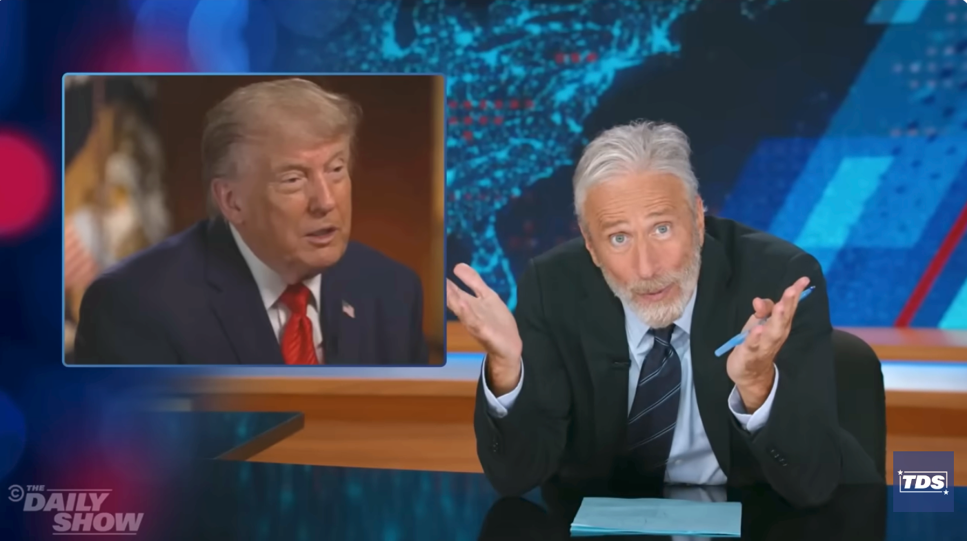 TV host gestures while discussing a news segment featuring a political figure on screen. Set is branded with "The Daily Show" and "TDS."