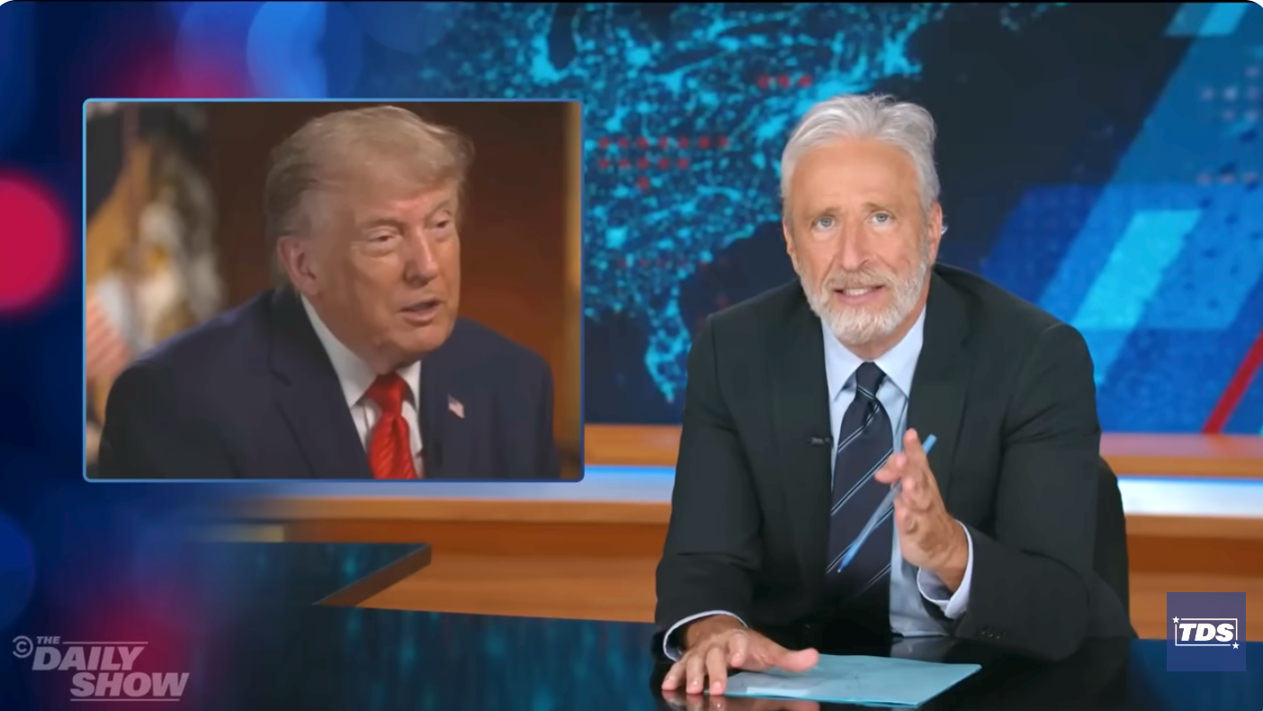 Man at news desk gestures while speaking, with a screen showing another man in a suit during a televised segment