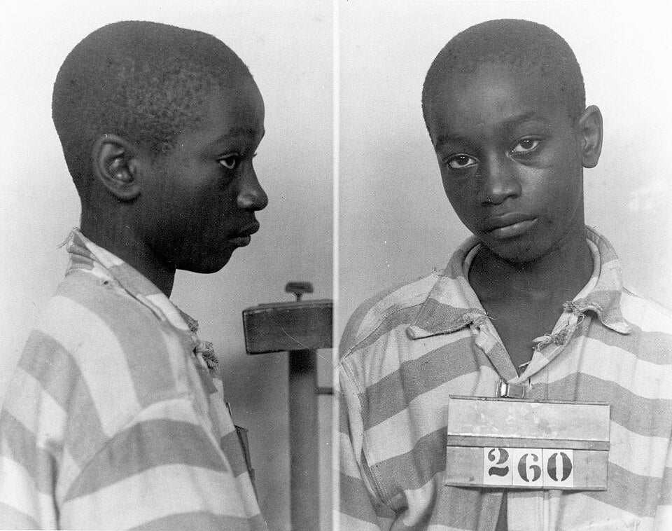A young idiosyncratic   successful  striped situation  attire, holding a numbered placard for a mugshot. They look  somber and somewhat  turned to the side