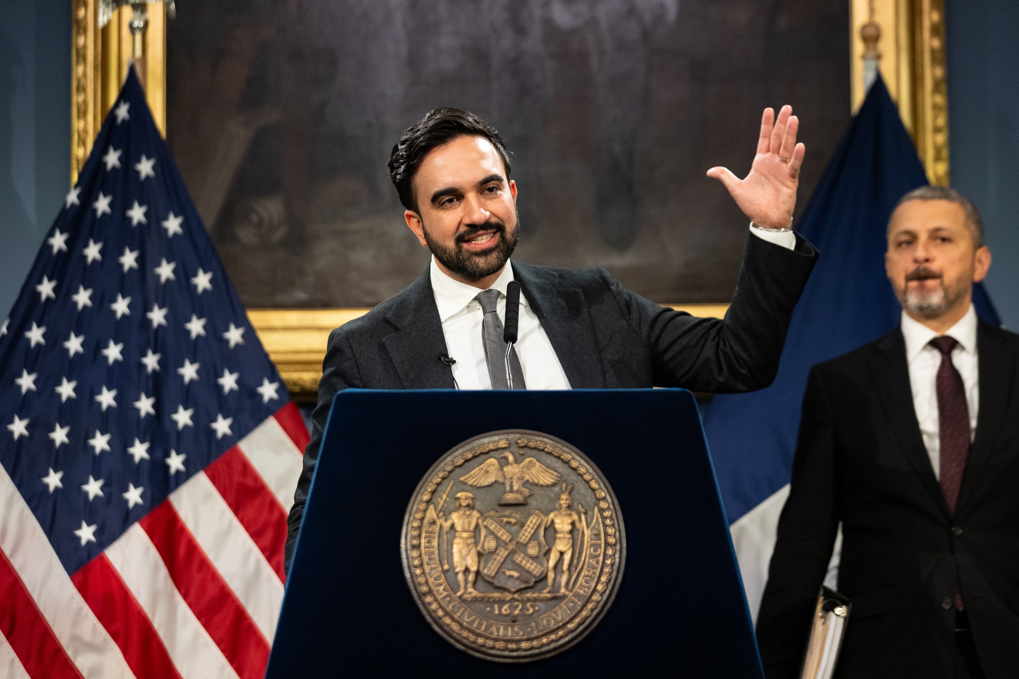 A man in a suit speaks at a podium with a government seal, raising one hand. An American flag is visible in the background