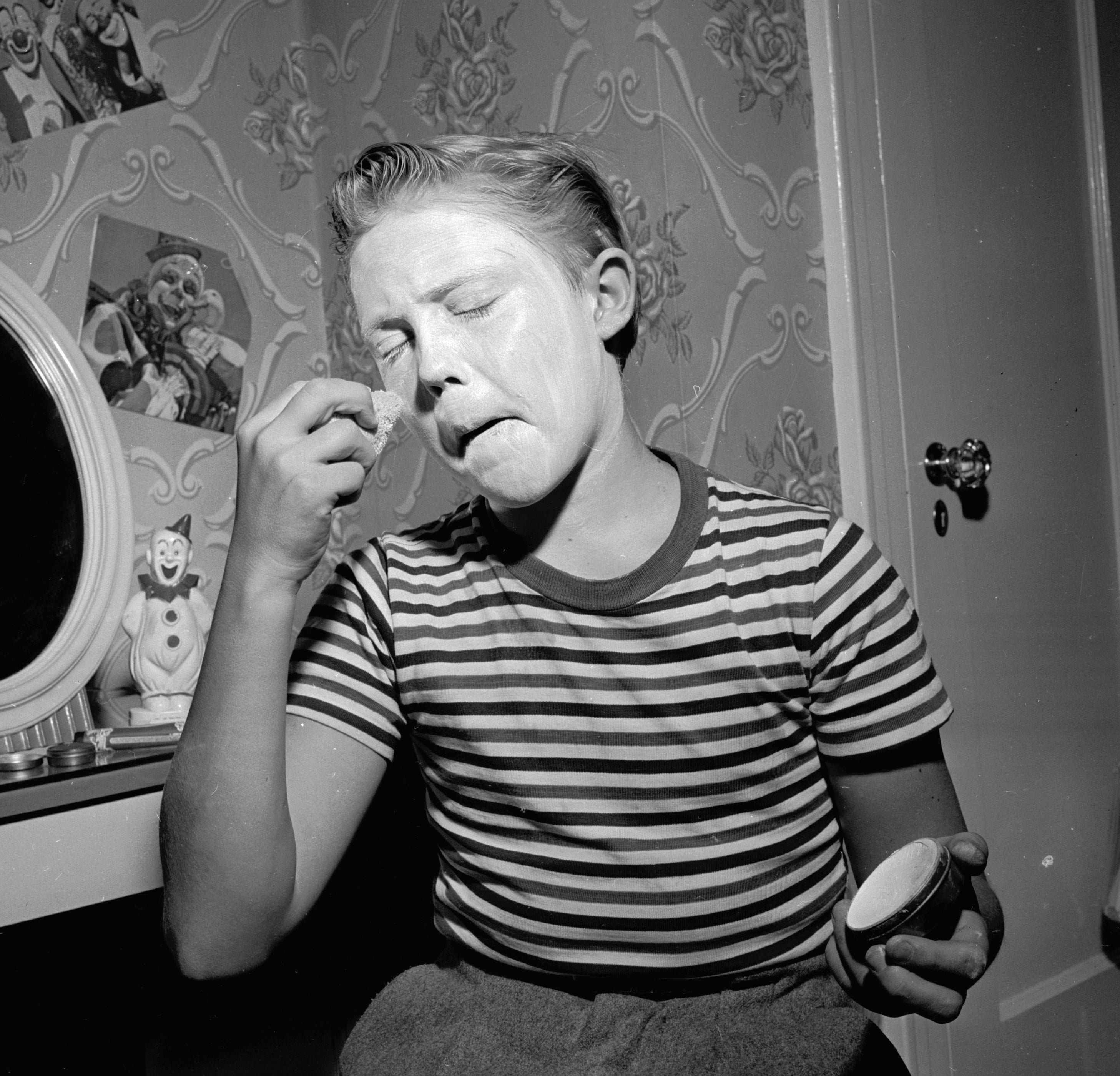 Christopher Walekn in a striped shirt applies face paint in front of a mirror, practicing clown makeup in a decorated room