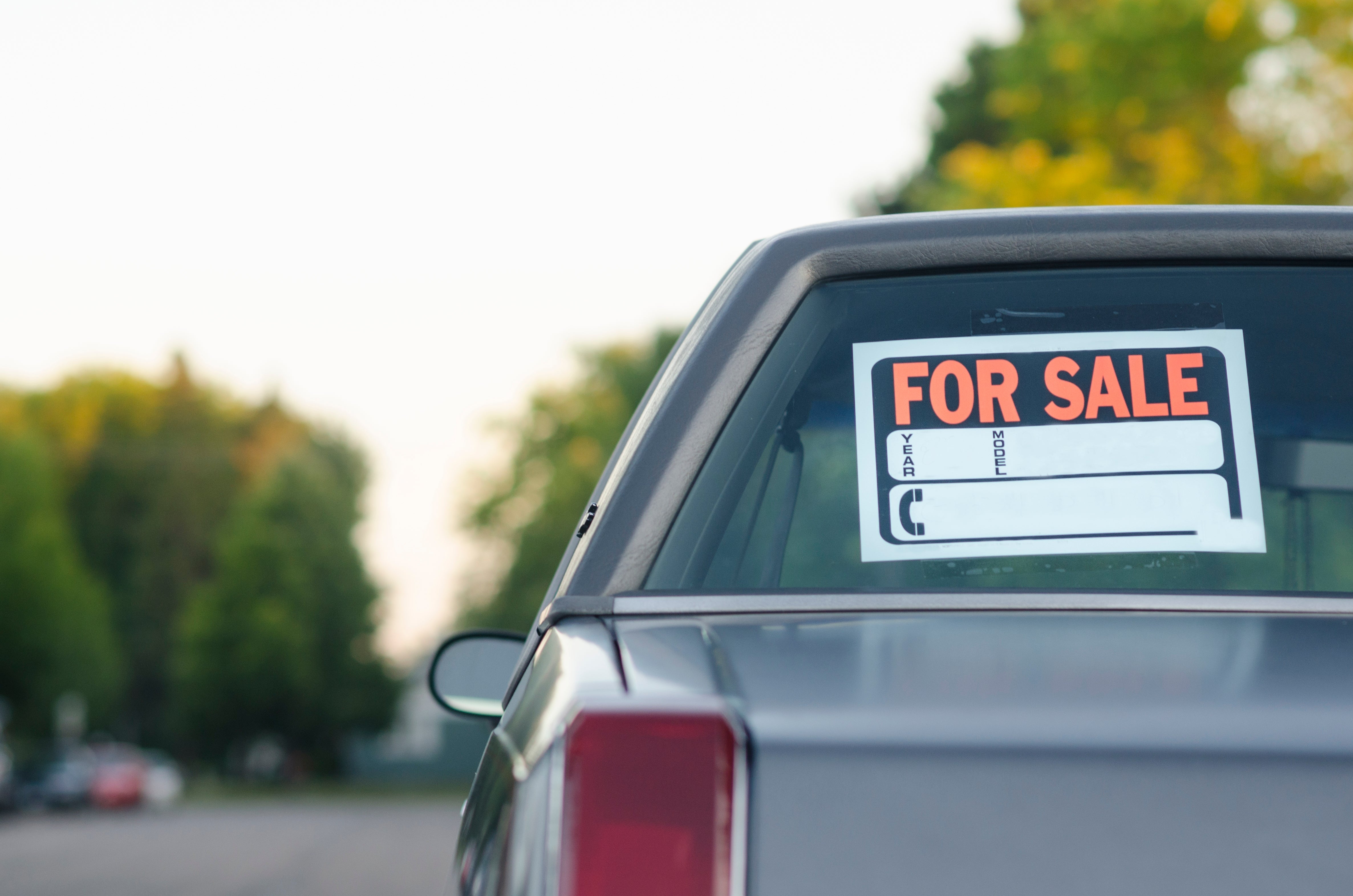 Car with "For Sale" sign in rear window on a suburban street. Sign lists year, model, and contact details. Trees in the background