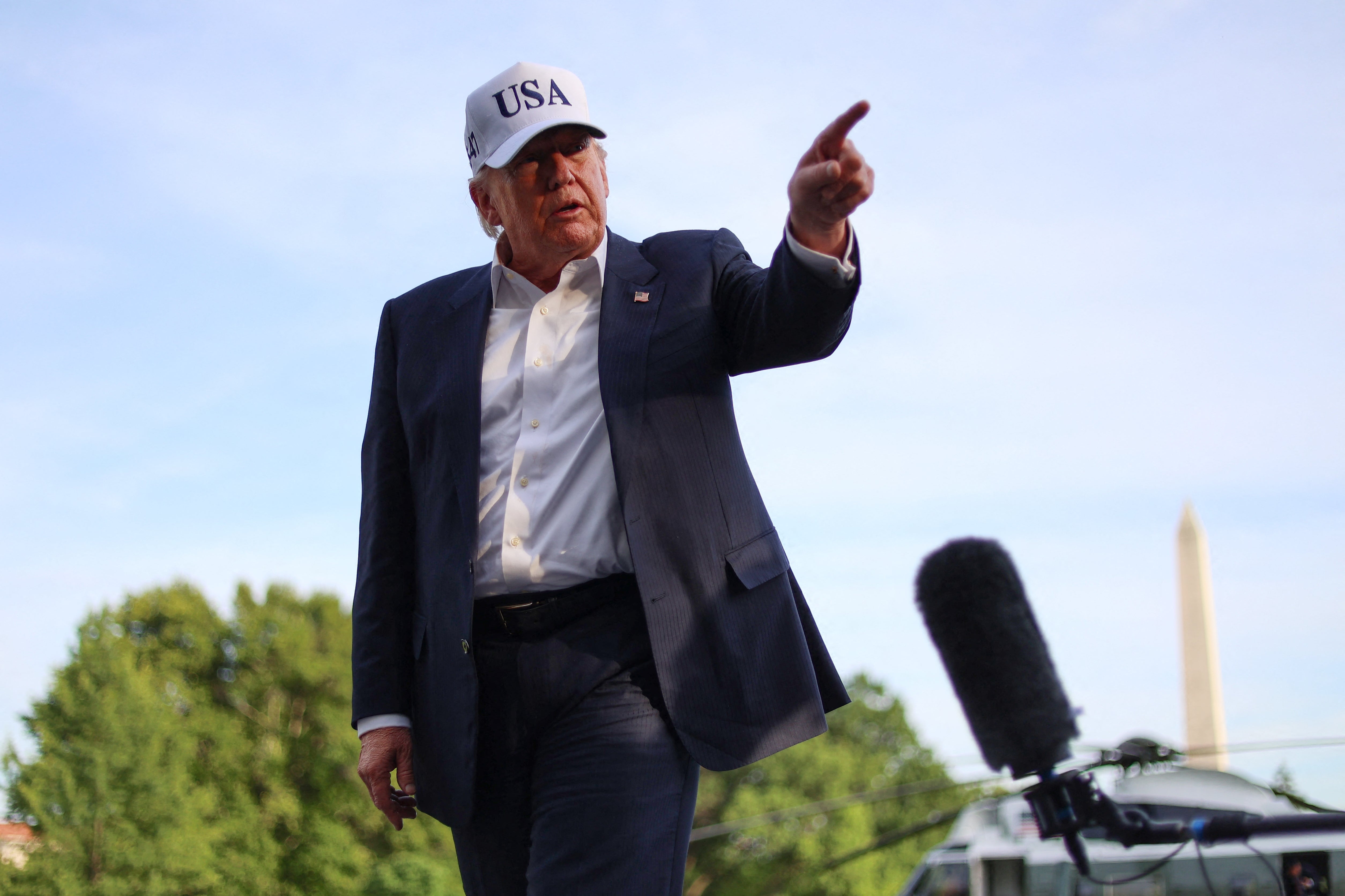 Person in a suit and USA cap gestures, standing outdoors with a microphone nearby, trees and an obelisk in the background