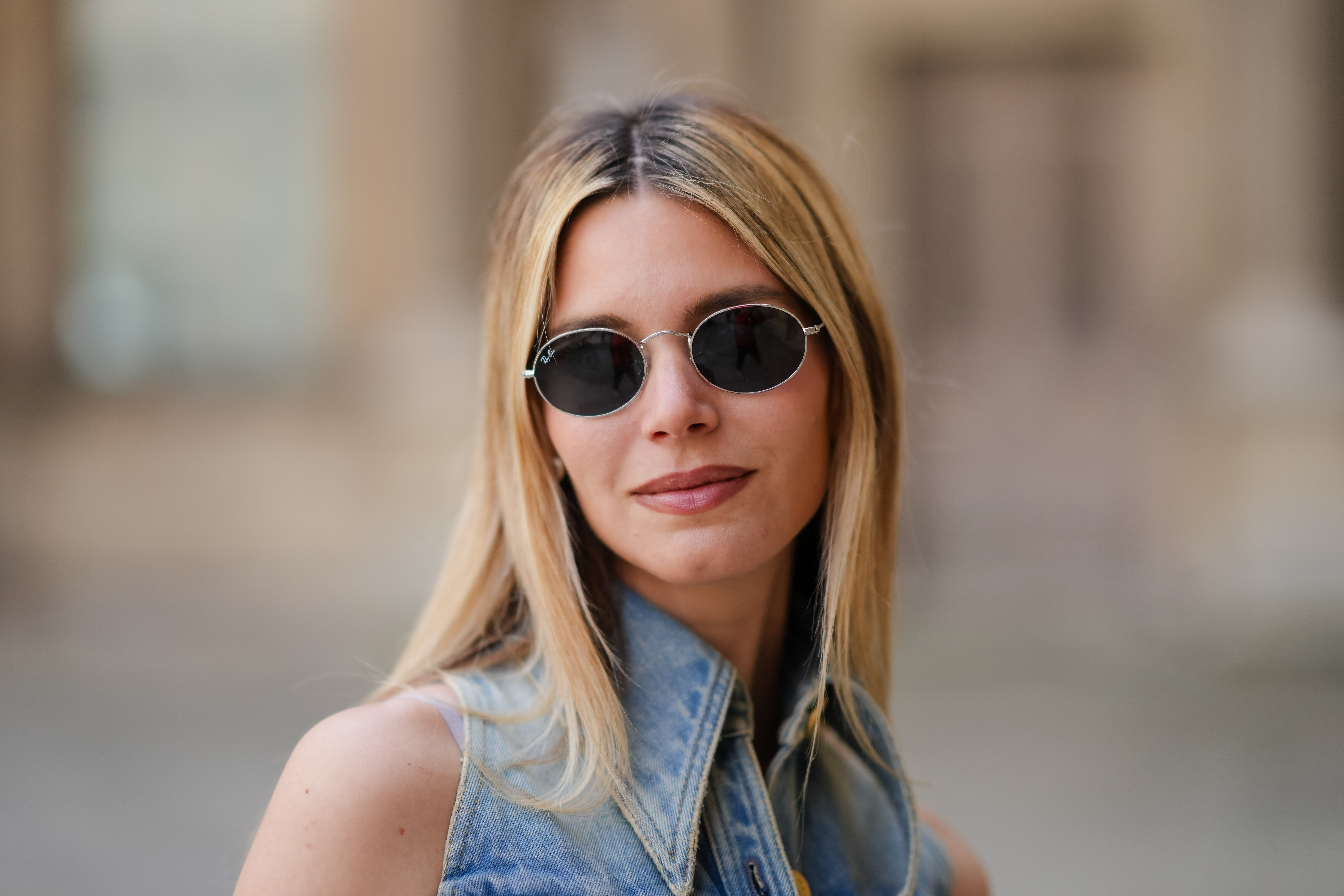 Woman with long hair wearing sunglasses and a denim vest, looking confidently at the camera