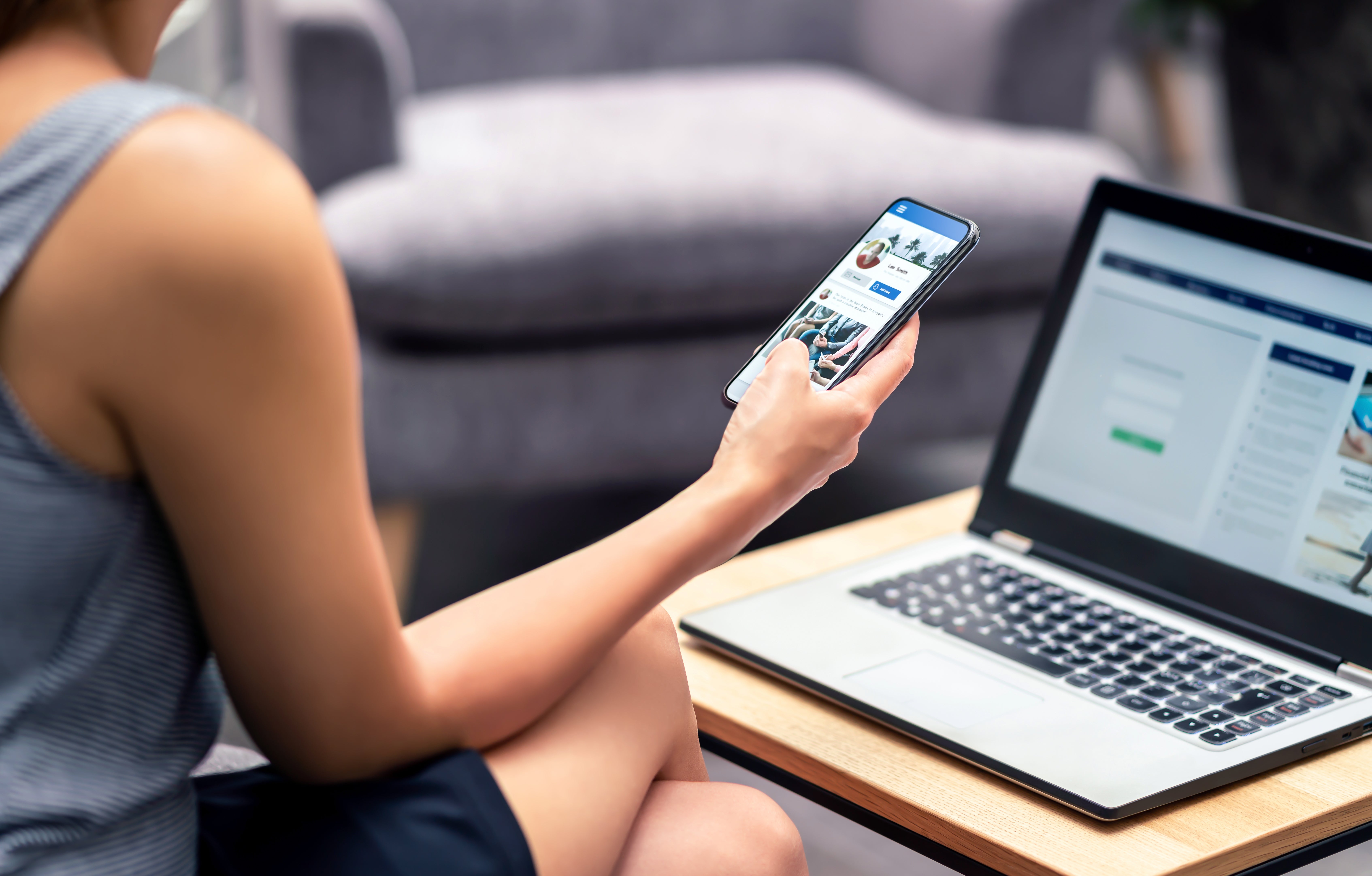 Person sitting and using a smartphone with a social media app, laptop open on a table with a login page visible