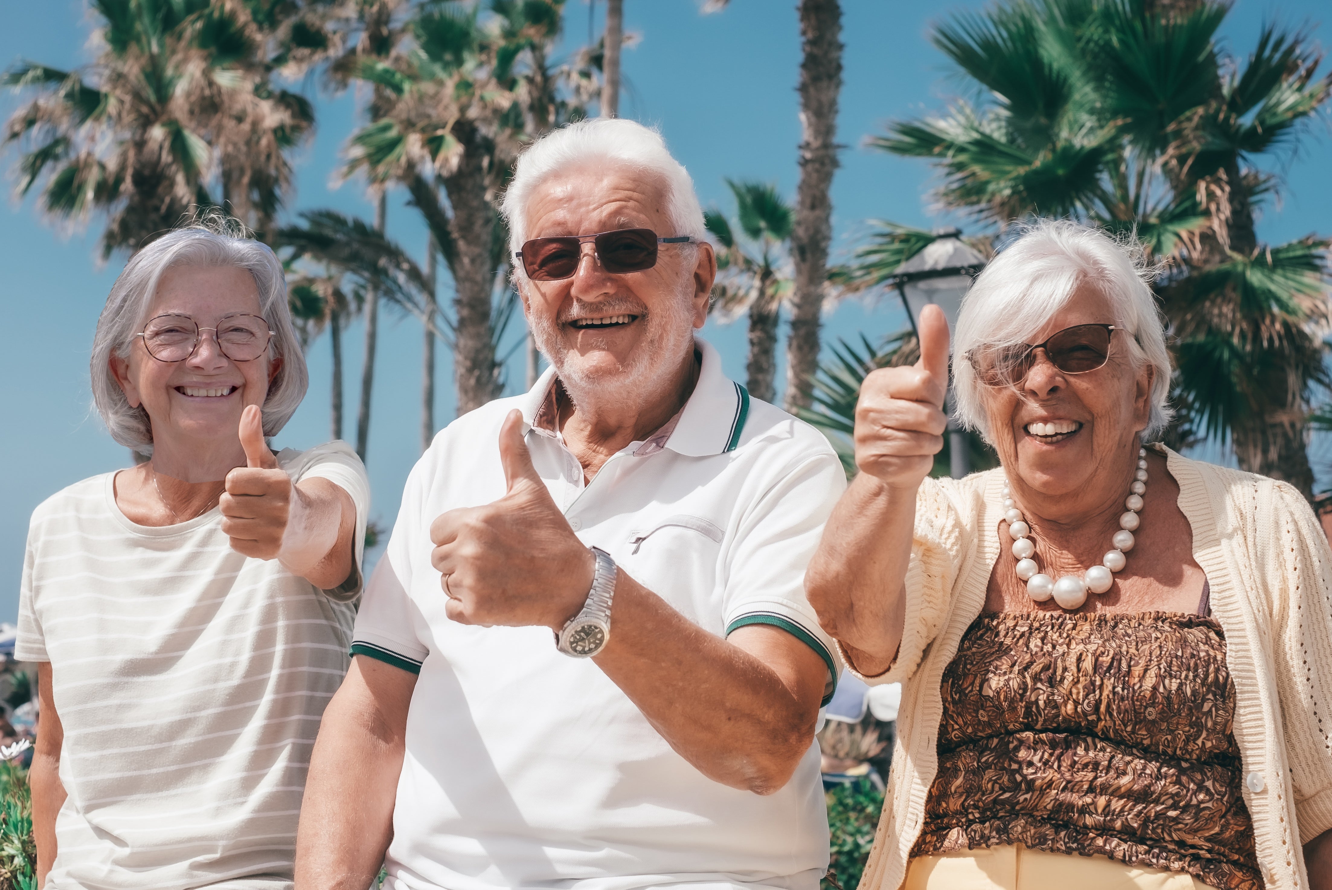 Three smiling older adults giving thumbs up, sitting outdoors with palm trees in the background