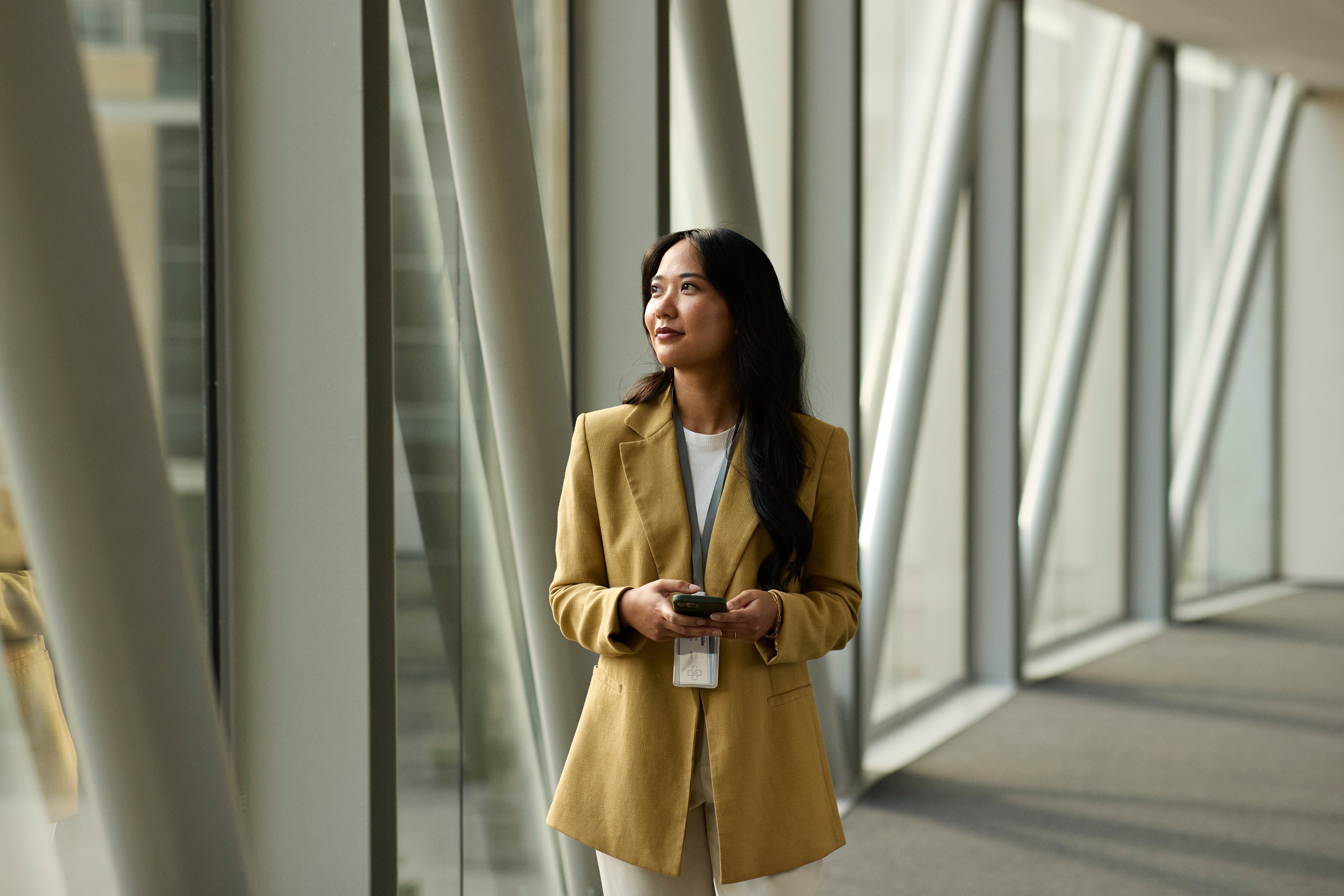 A person stands in a modern corridor, wearing a tailored suit and holding a phone, looking thoughtfully out of a window