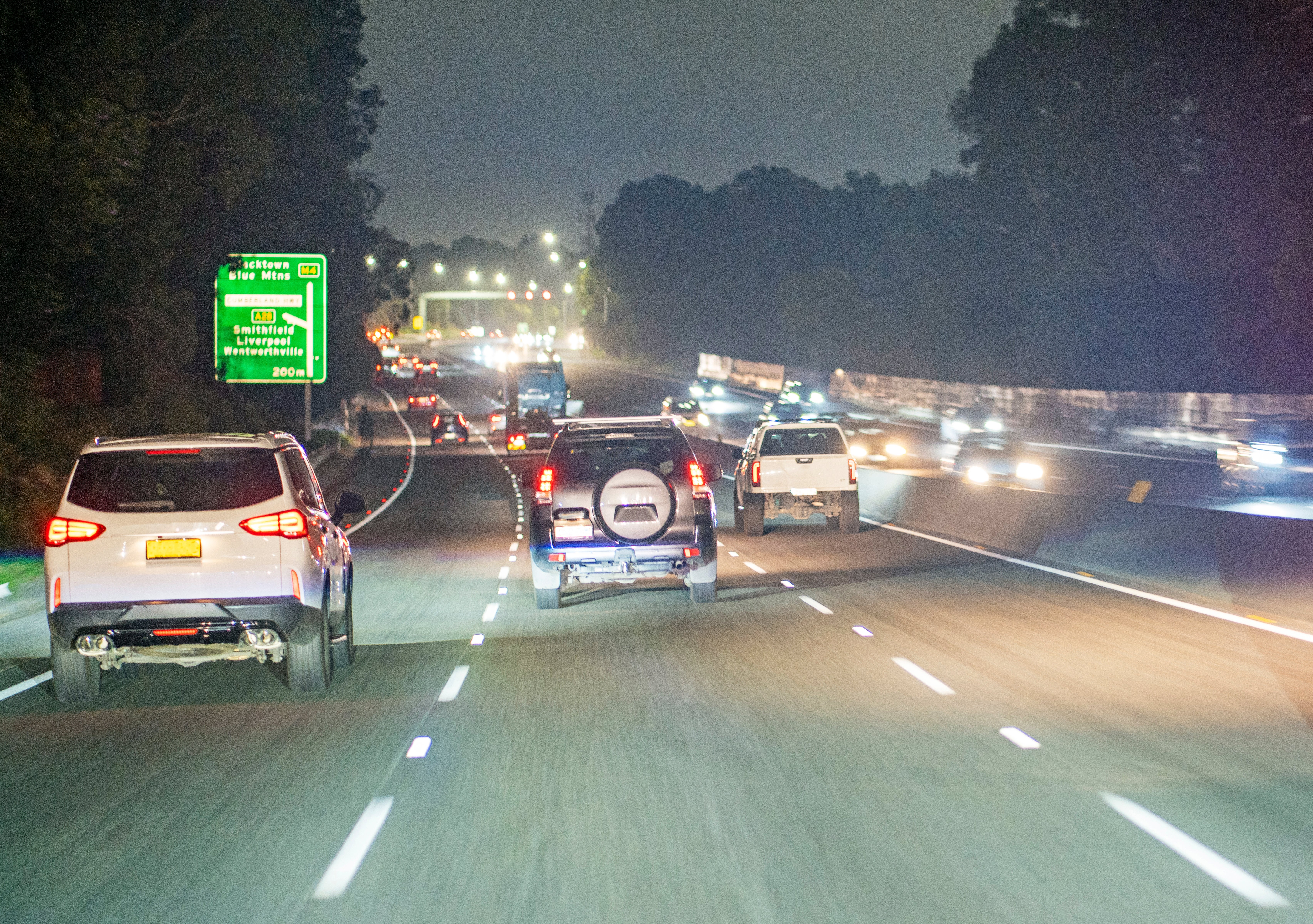 Cars driving on a multi-lane highway at night, with trees lining the road and illuminated road signs visible