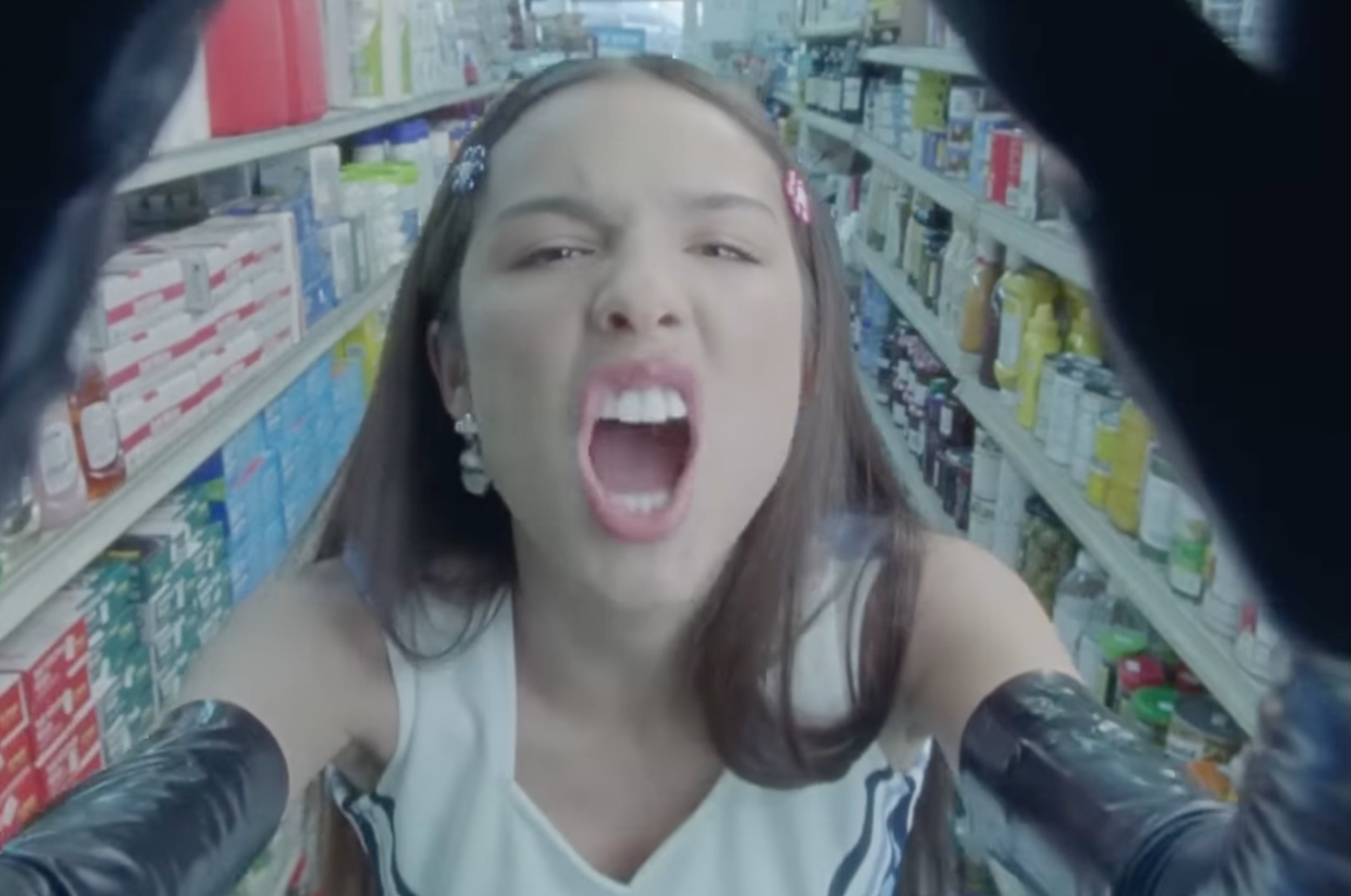 Person in a supermarket aisle with an expressive face, reaching toward the camera, wearing a sleeveless top and long gloves