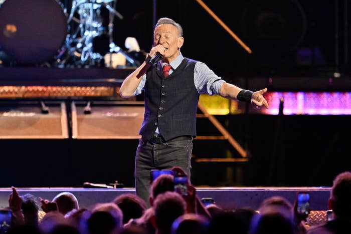 A singer performs energetically on stage, wearing a vest and tie, holding a microphone to the crowd's delight