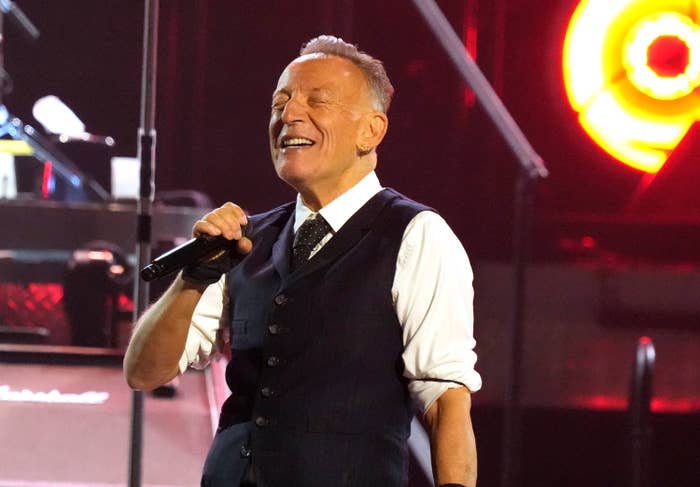 A person energetically singing on stage, holding a microphone, wearing a vest over a shirt and tie, with musical equipment in the background