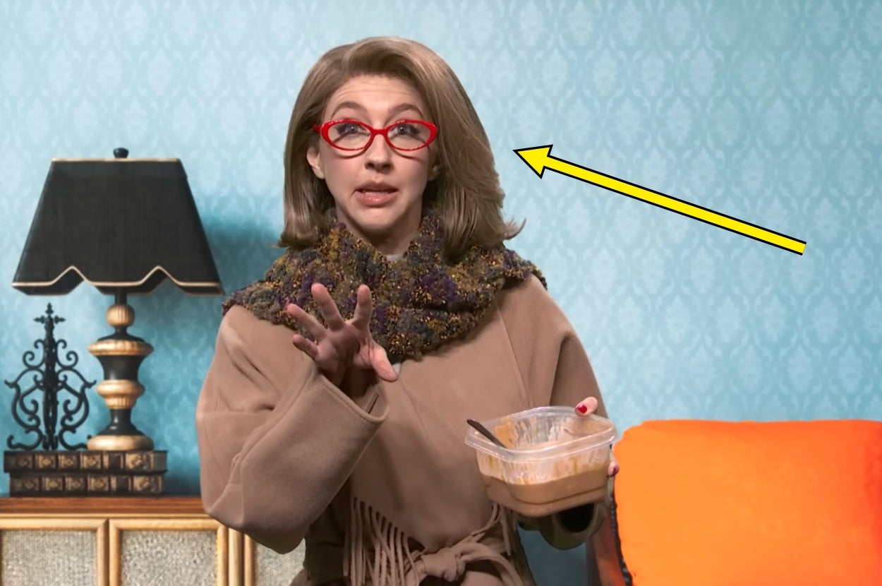 Person wearing a bulky scarf and red glasses holds a container while gesturing. They are in a retro-styled room with a lamp and orange cushion
