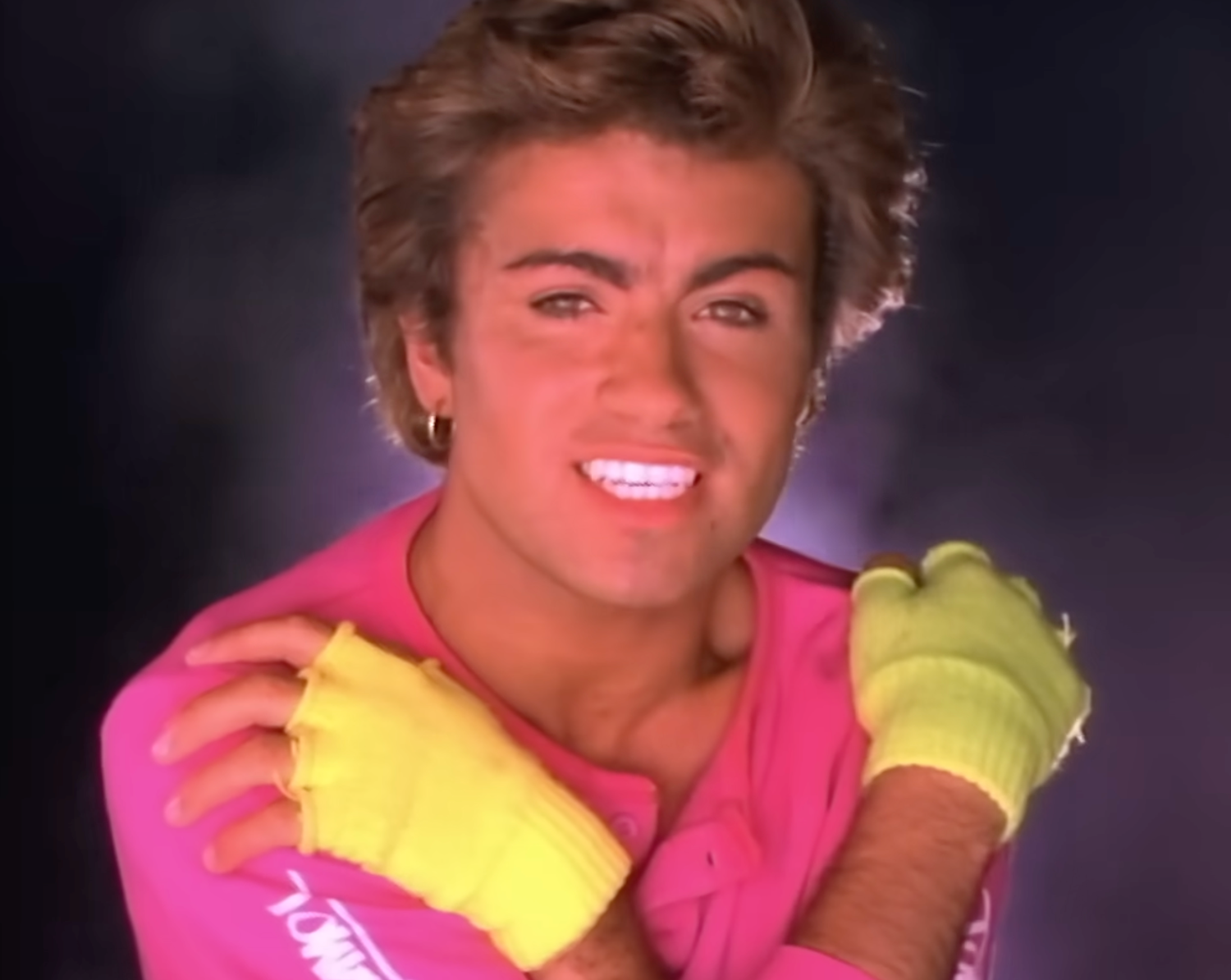 Musician smiling with styled hair, wearing a pink top and bright gloves, posing with crossed arms