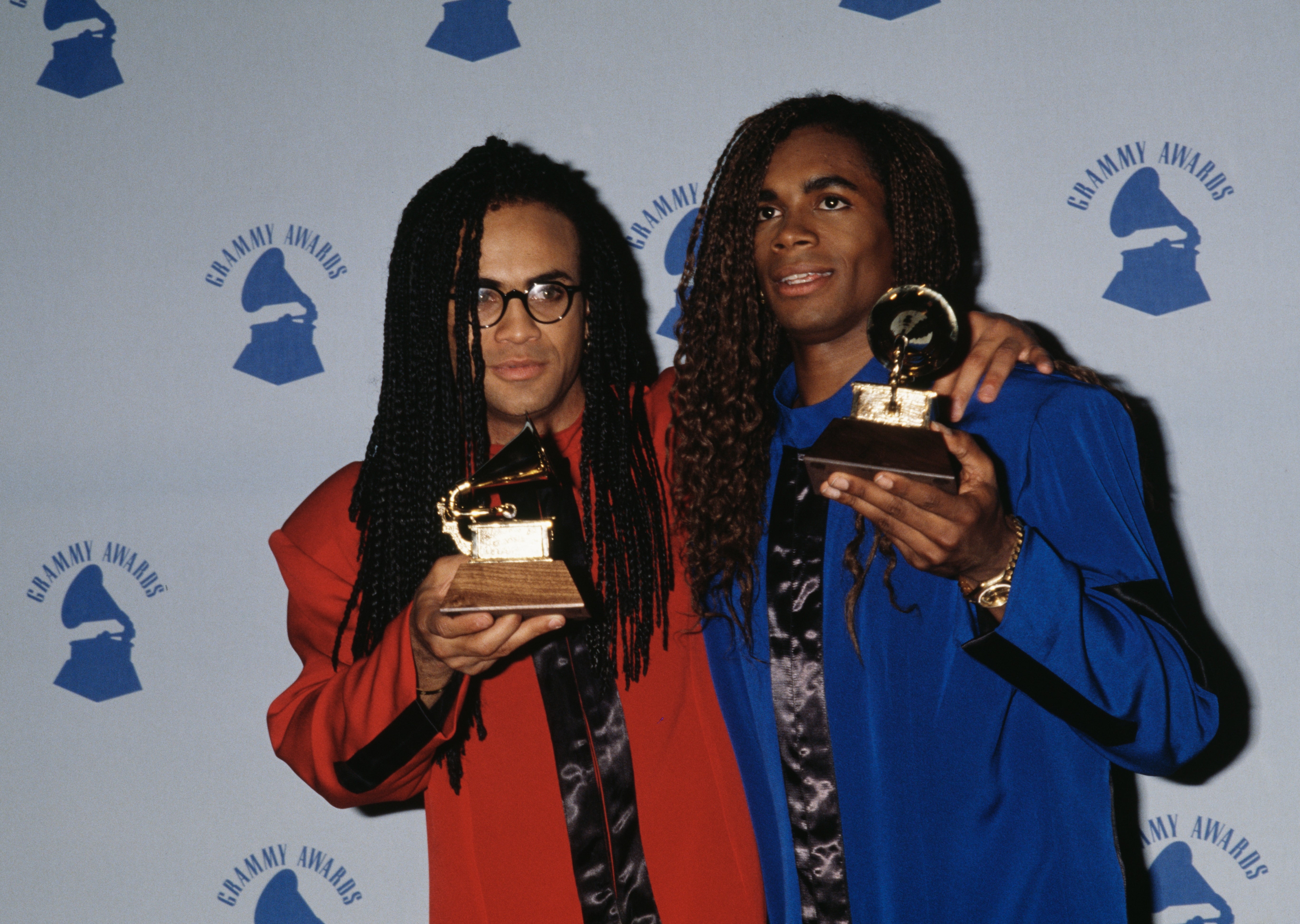 Dua orang memegang penghargaan Grammy, mengenakan setelan tebal dengan bahu berlebihan, berdiri di depan latar belakang berlogo Grammy
