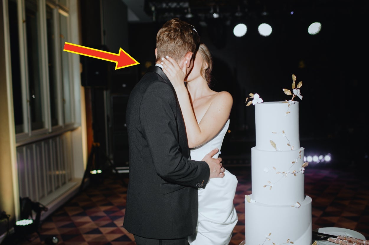 A couple shares a romantic kiss next to a tiered wedding cake, while dressed in formal attire at a celebration