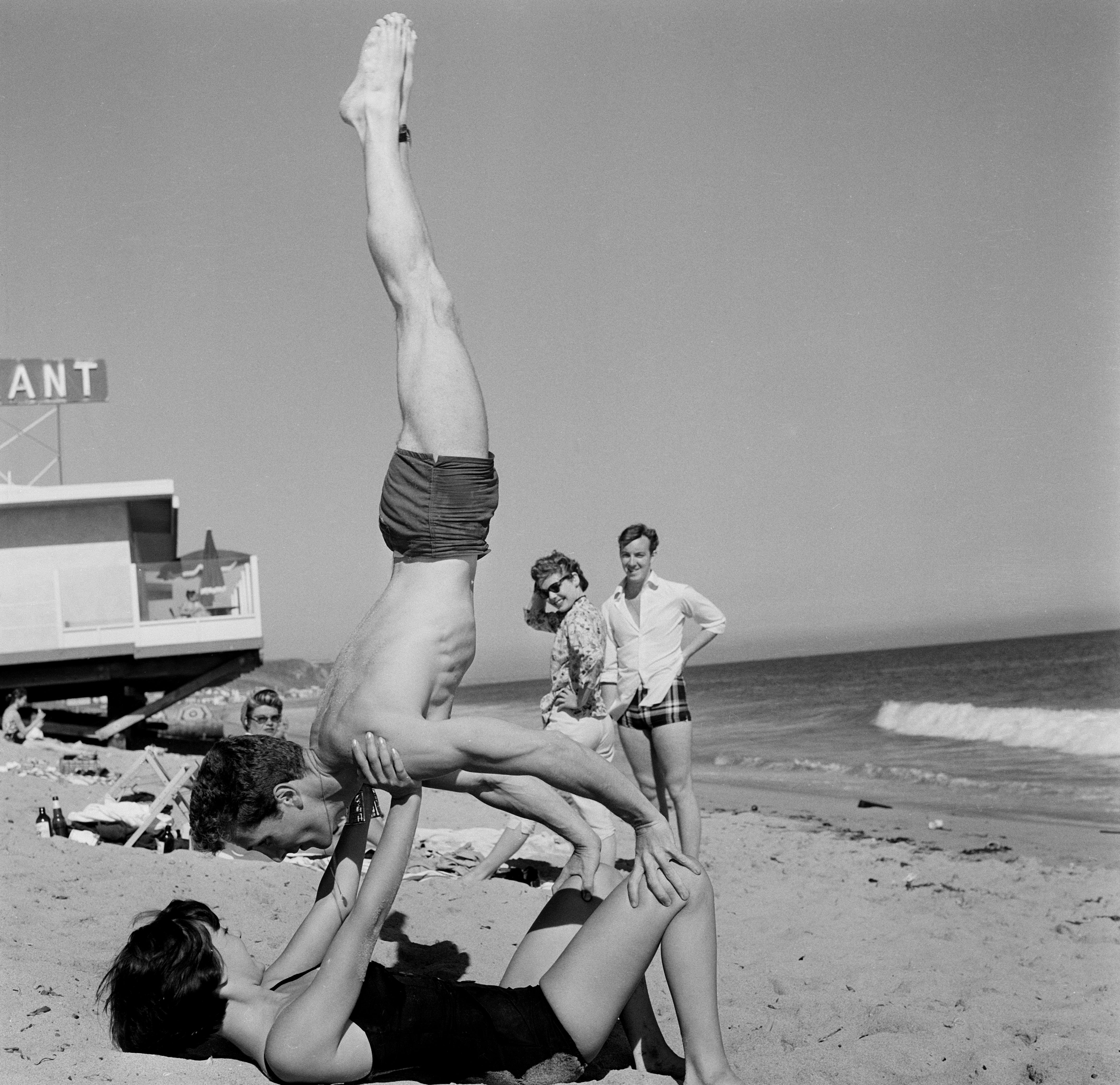 Steve Rowland and Natalie Wood perform an acrobatic balance on a beach, with his hands on her knees and her hands on his shoulders, surrounded by onlookers