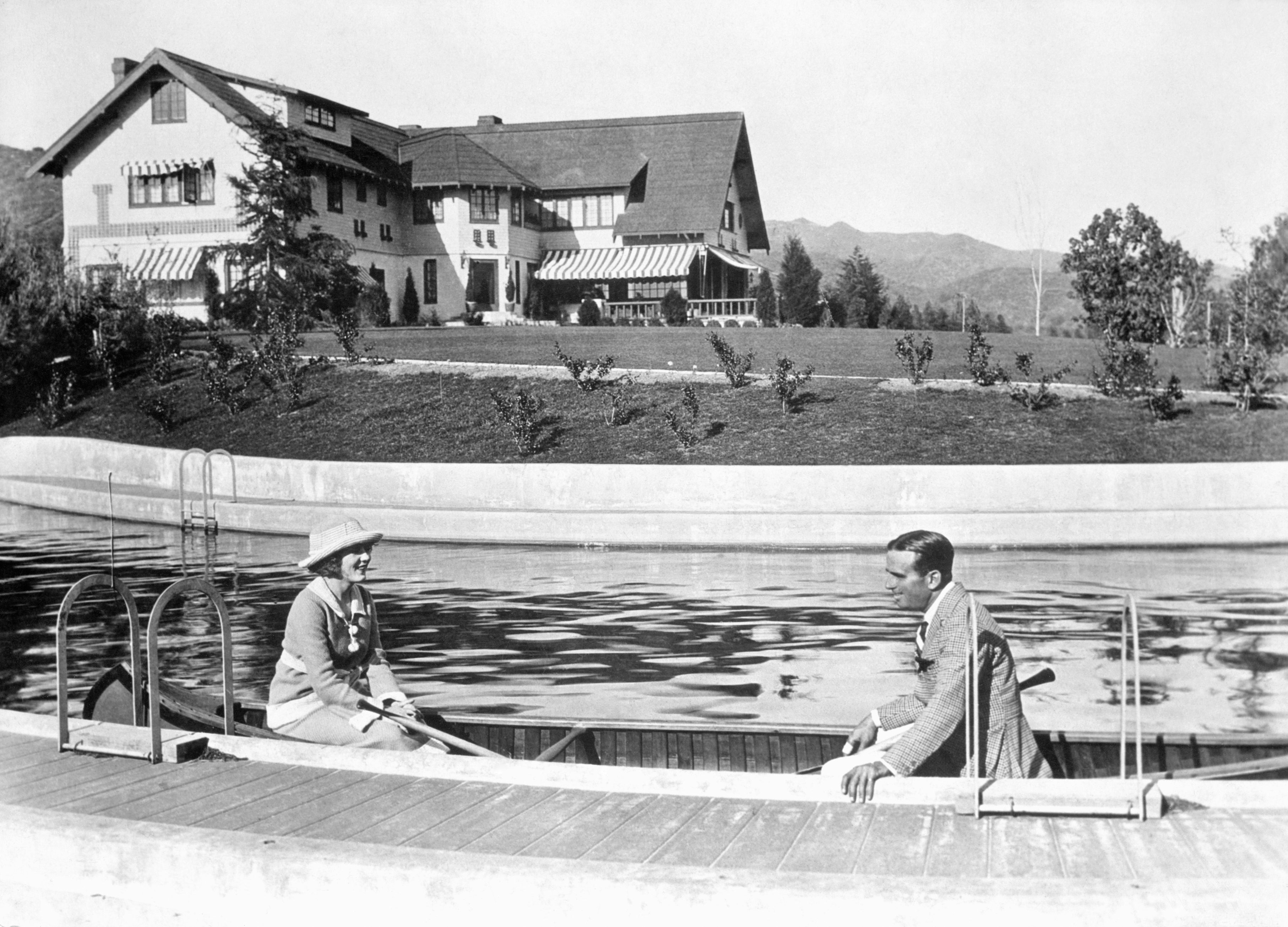 Douglas Fairbanks and Mary Pickford in a canoe on a pool near a large house.