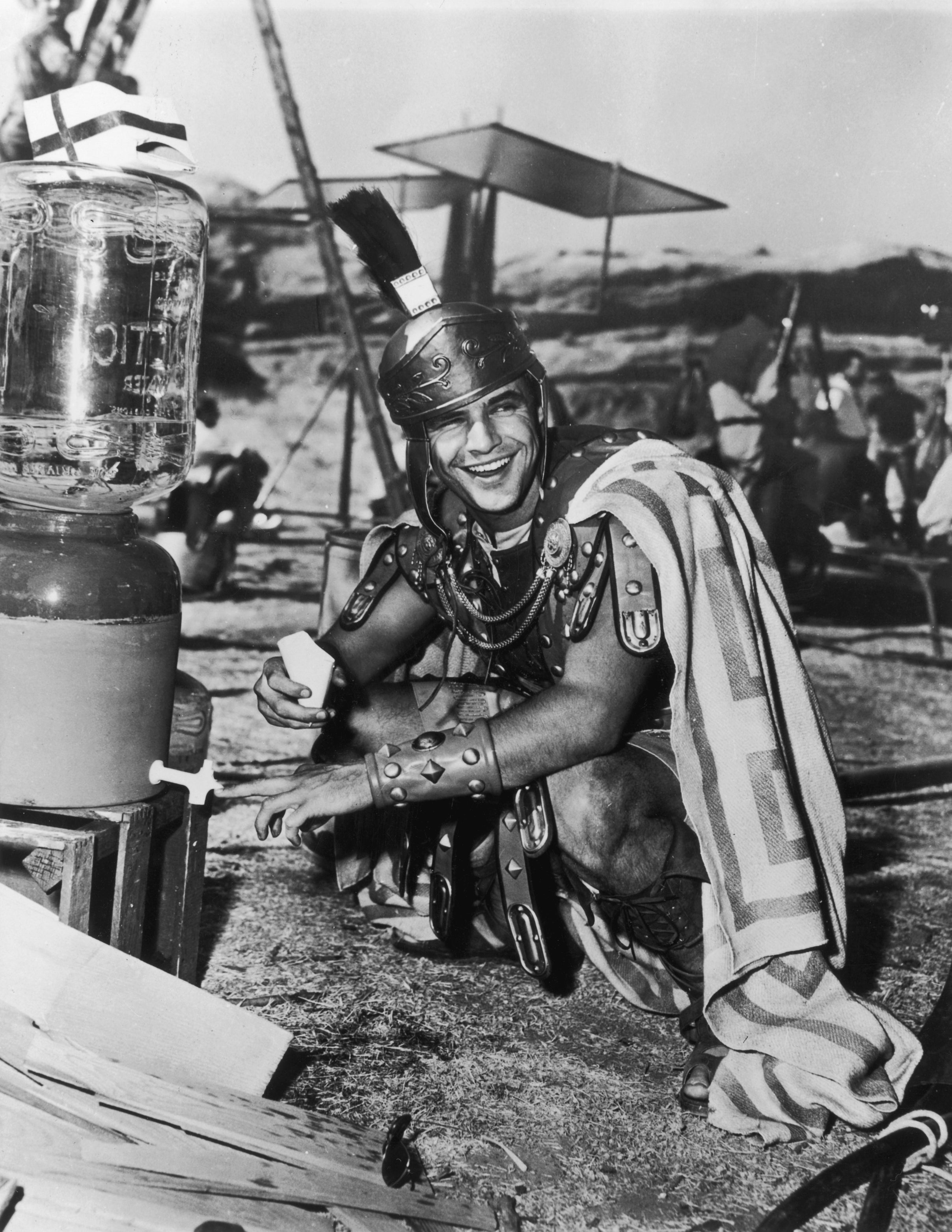 Marlon Brando in Roman gladiator costume crouching and laughing while holding a cup beside a large water cooler on a film set