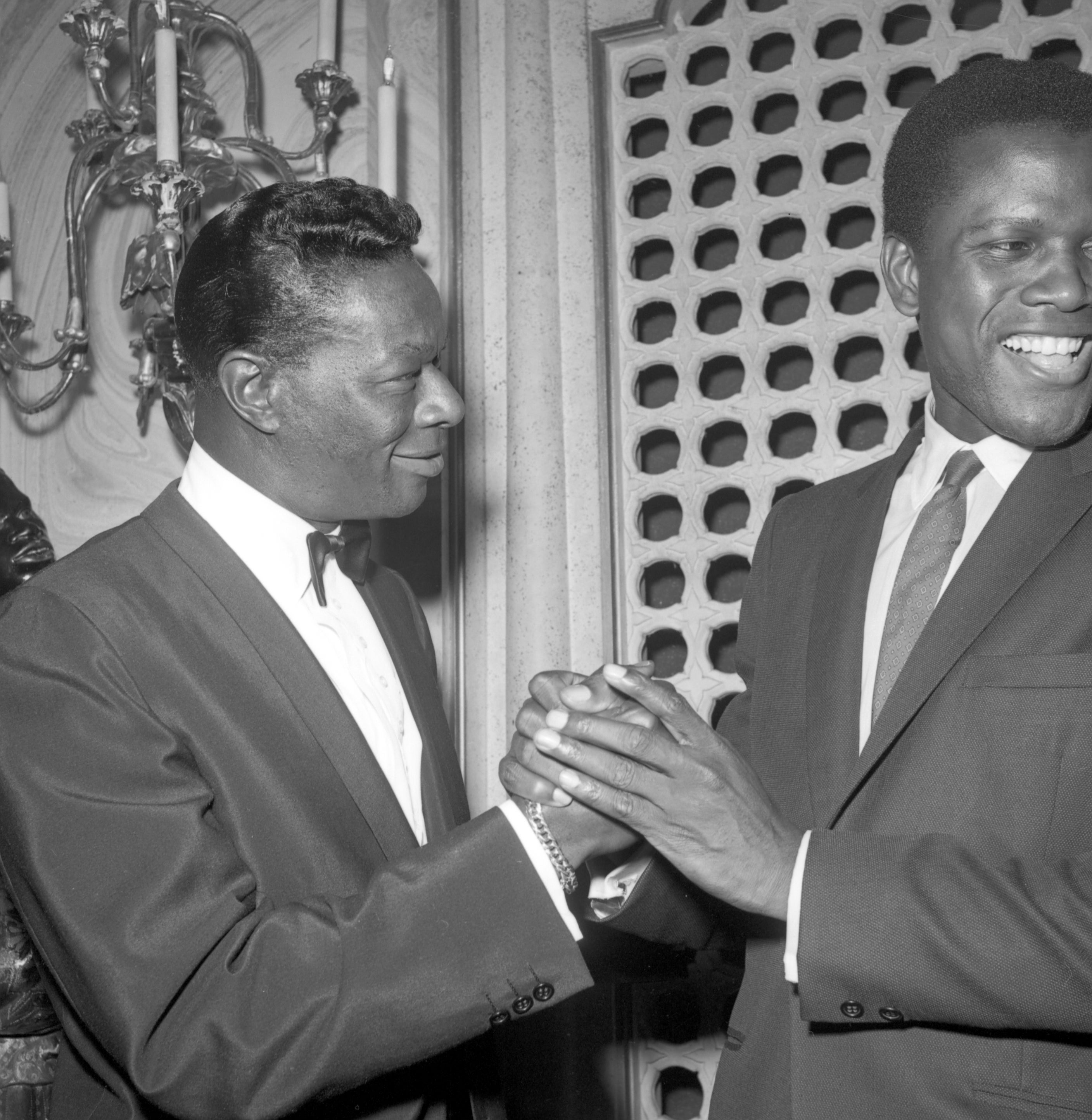 Nat King Cole and Sidney Poitier are shaking hands and smiling, standing indoors