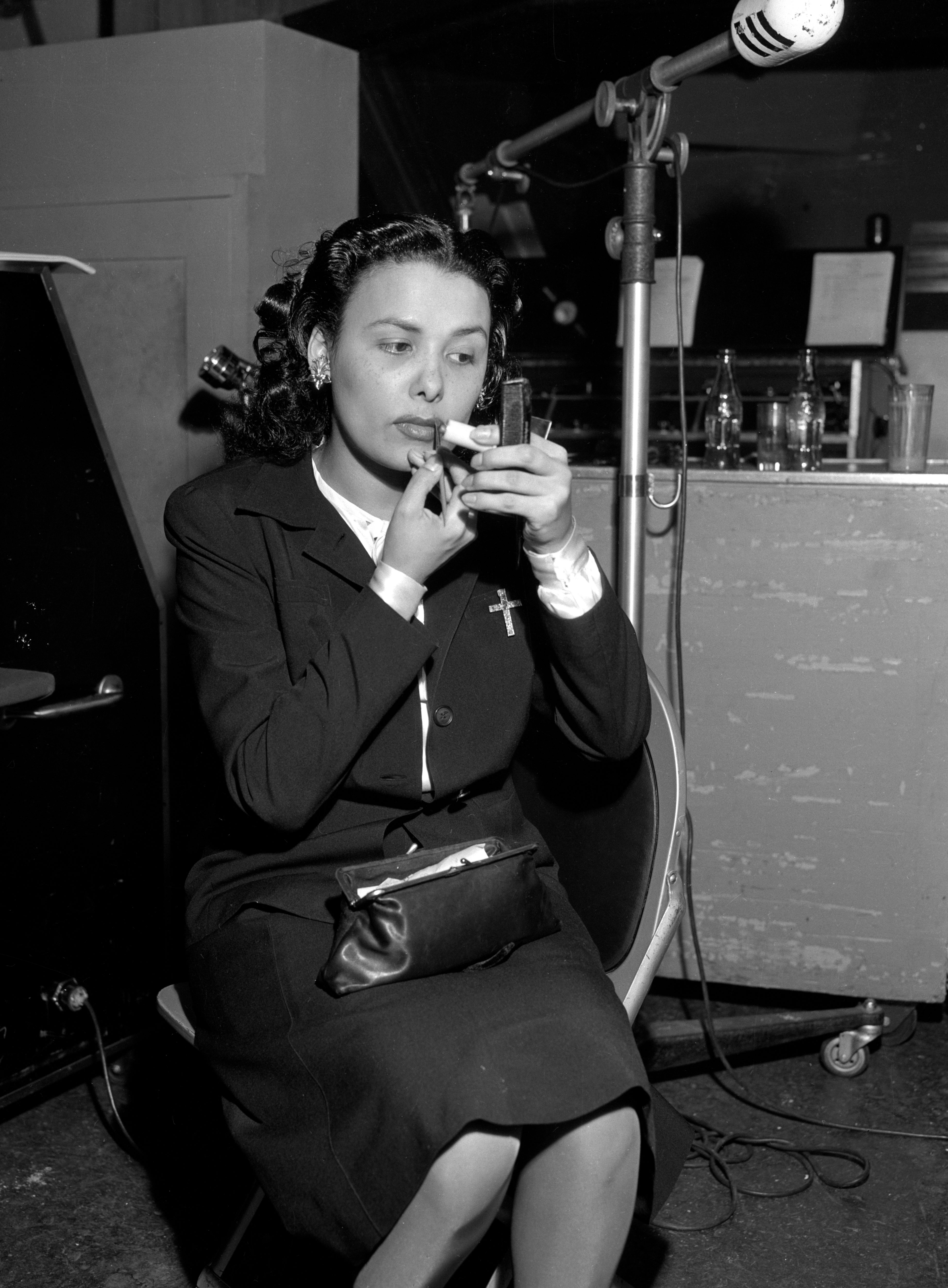 Lena Horne in a tailored suit applies lipstick while seated backstage, holding a compact mirror. She has styled hair and a small purse on her lap