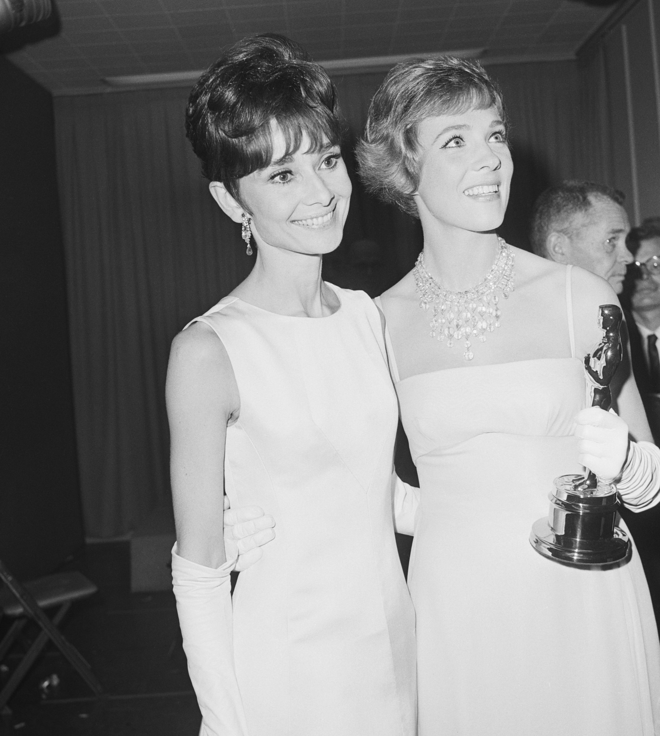 Audrey Hepburn and Julie Andrews in elegant, sleeveless gowns and gloves stand together, with Julie Andrews holding an Academy Award statue