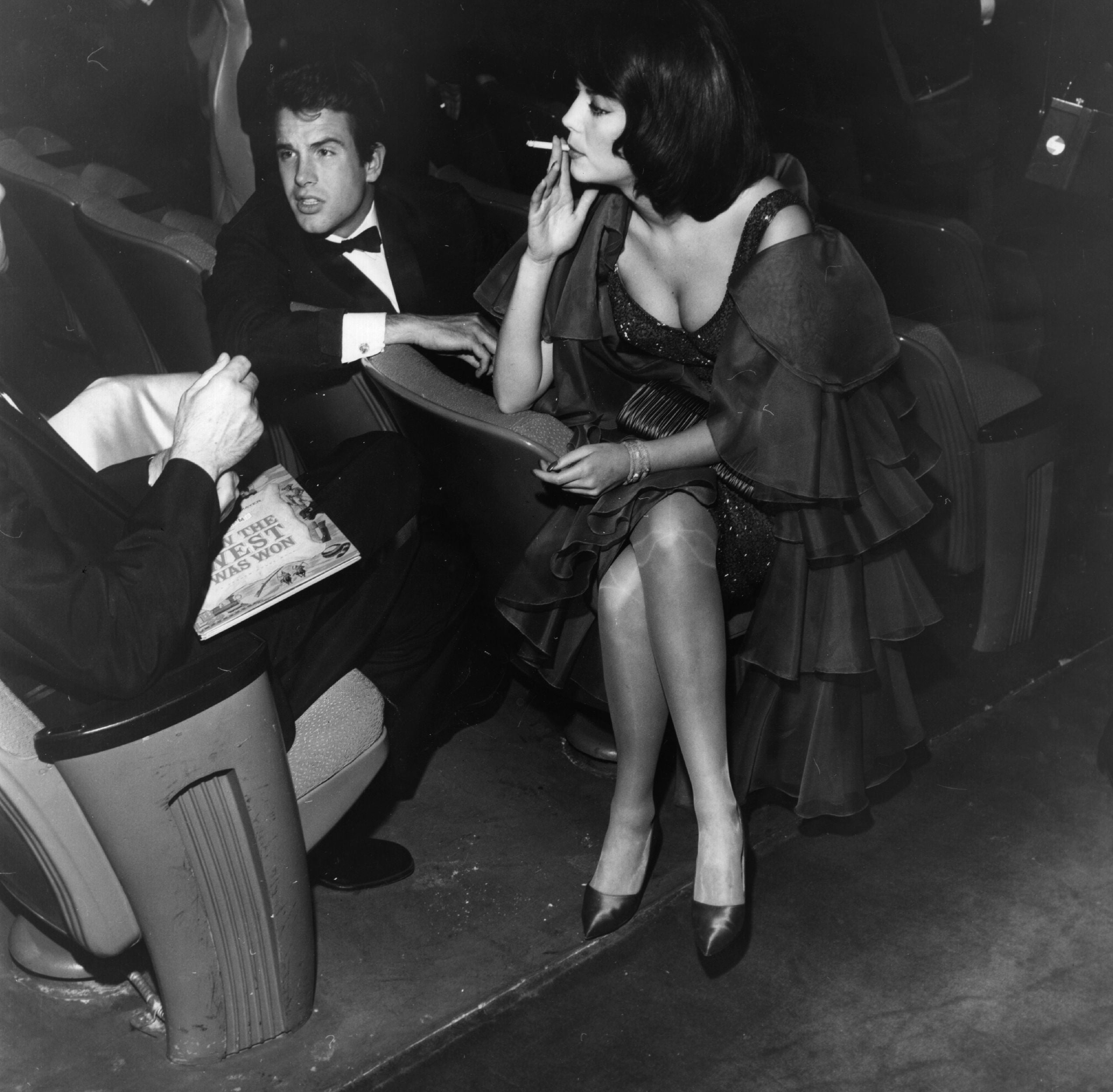 Warren Beatty in a tuxedo and Natalie Wood in an elegant, layered gown with a cigarette seated, conversing at a formal event