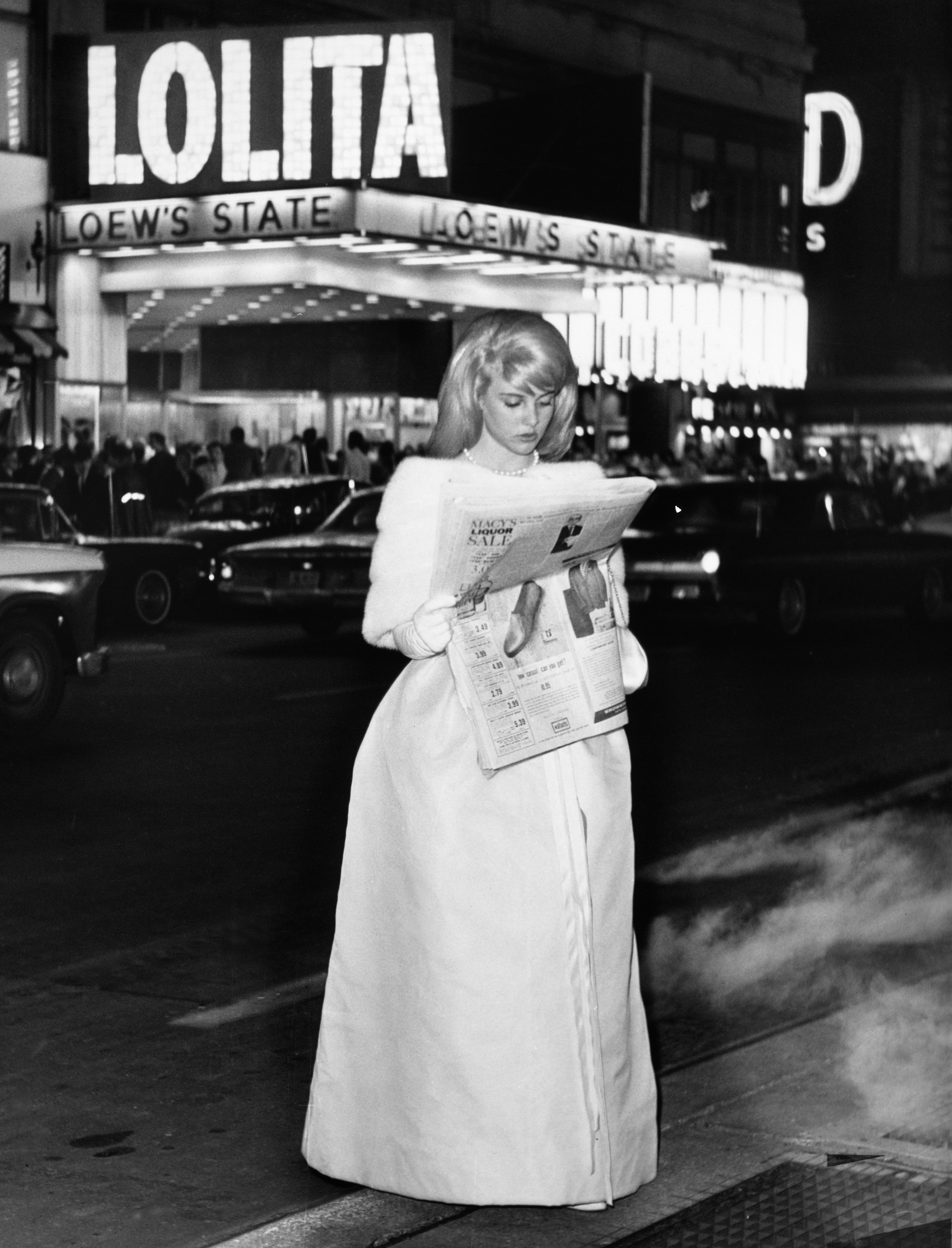 Sue Lyon in an elegant long coat reads a newspaper on a city street at night, with Loew's State theater and "Lolita" on the marquee in the background