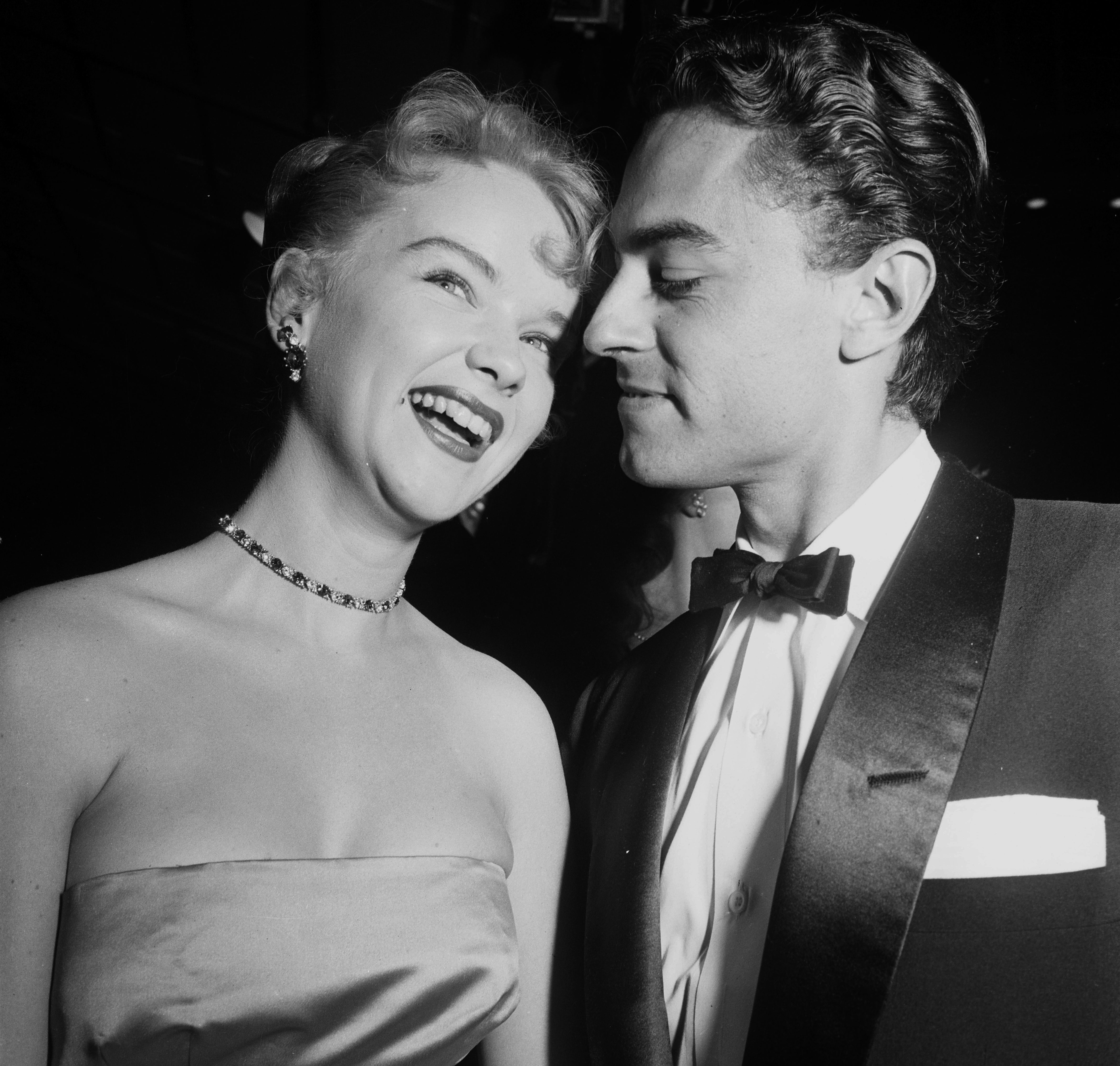 Anne Francis smiling  in a strapless gown with a choker necklace stands close to Bam Price in a tuxedo, captured in a candid moment