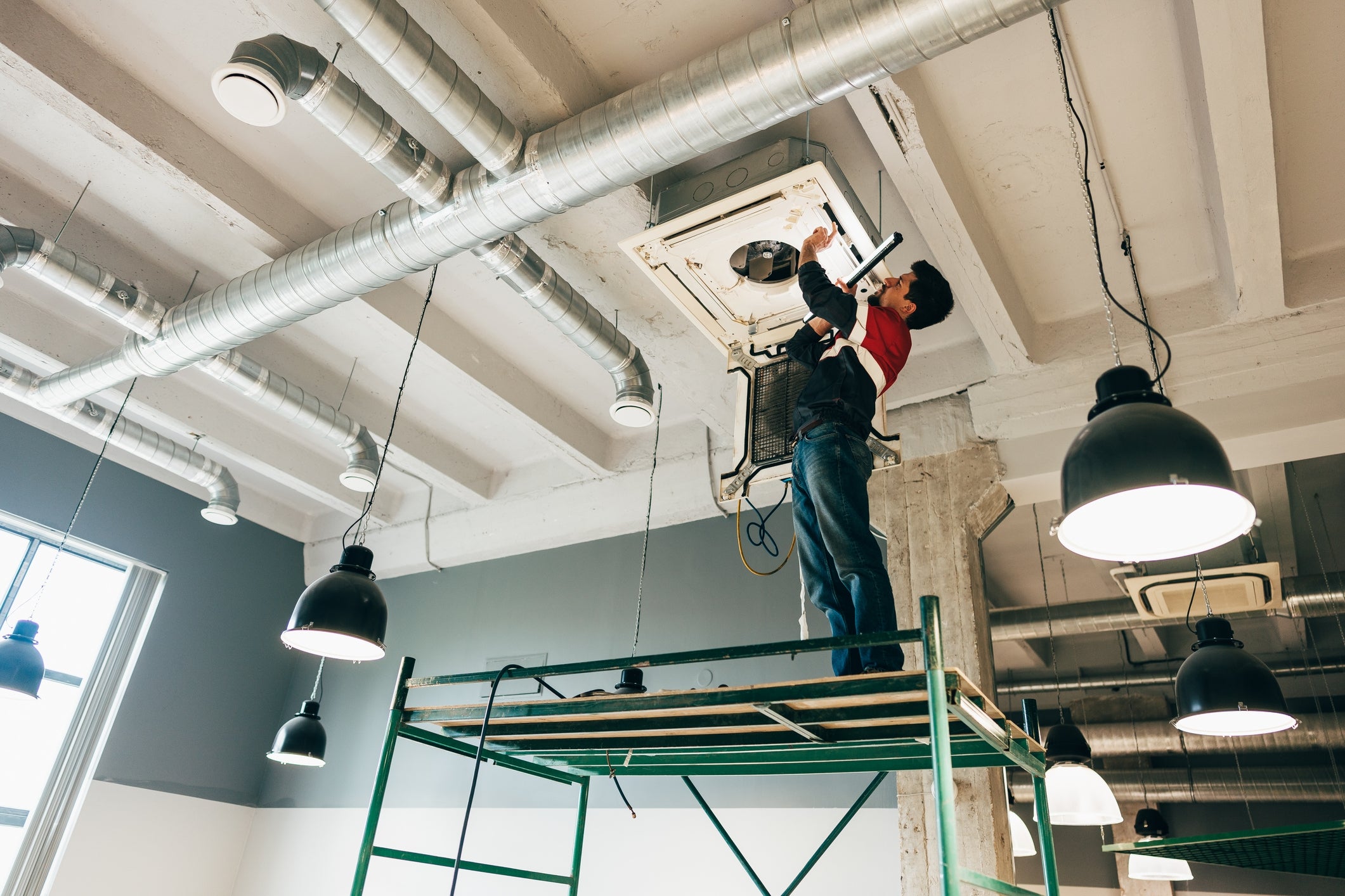 A worker stands on scaffolding, repairing a ceiling air conditioning unit in an industrial space