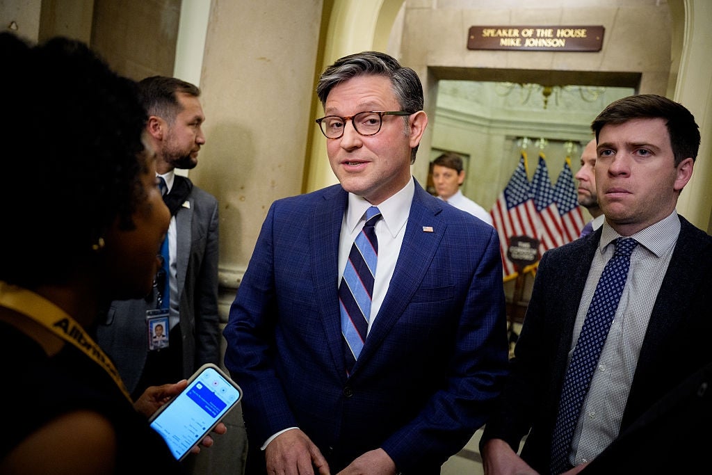 Two people conversing in a government building hallway, one holding a phone displaying information. Flags and a "Speaker of the House" sign in the background