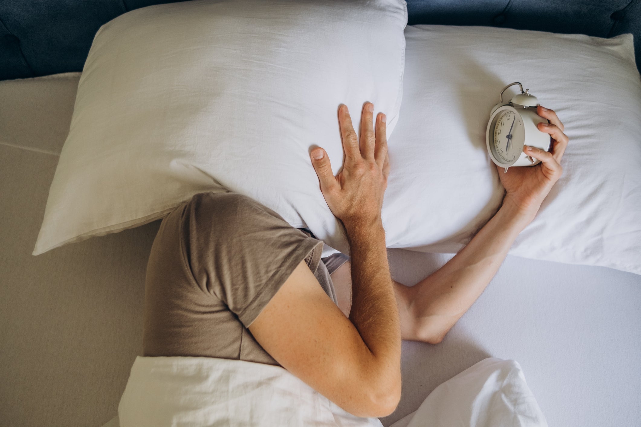 Person in bed hides face with a pillow while holding an alarm clock, conveying reluctance to wake up