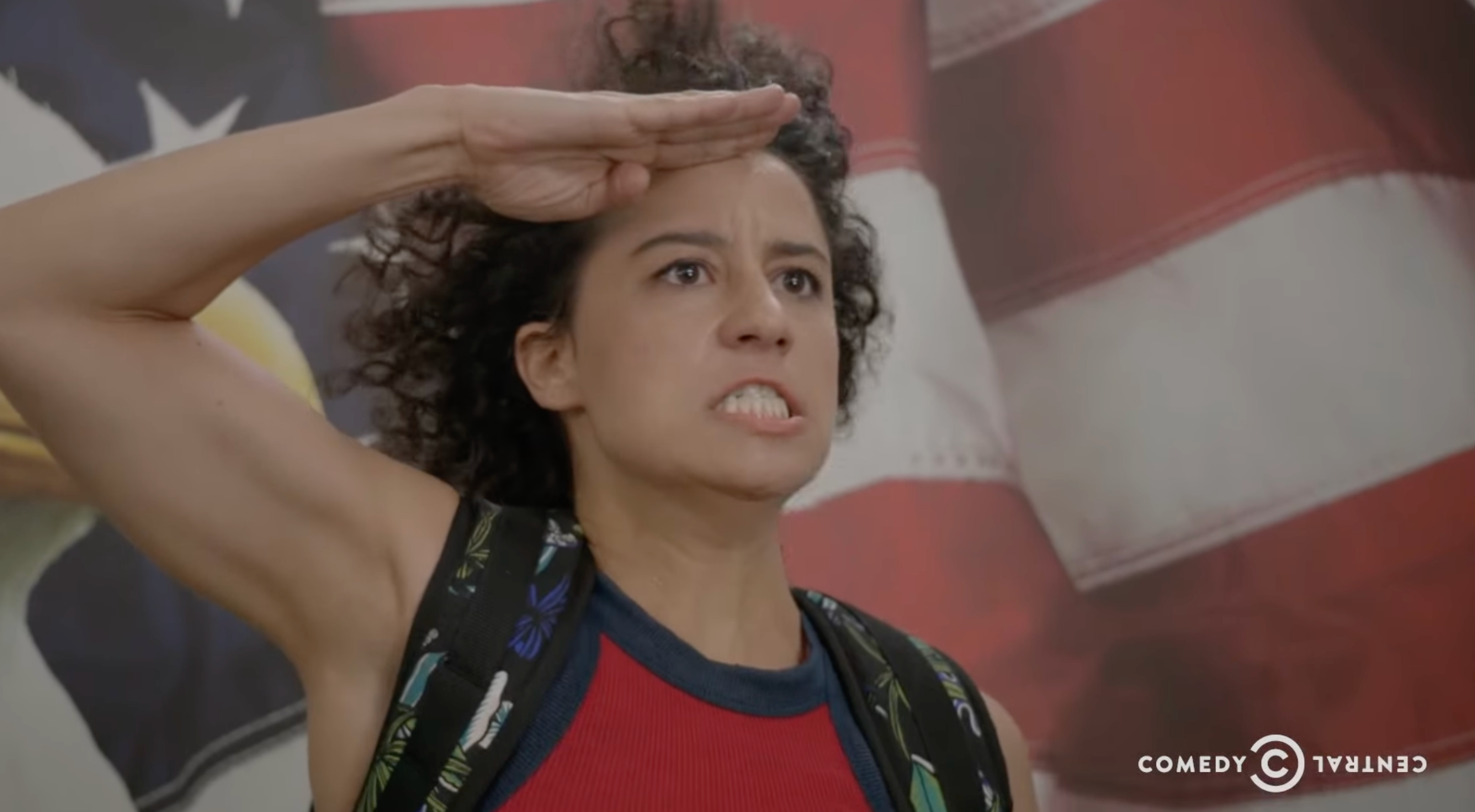 Person with curly hair saluting intensely in front of a large flag. They wear a backpack and a casual outfit. Comedy Central logo visible