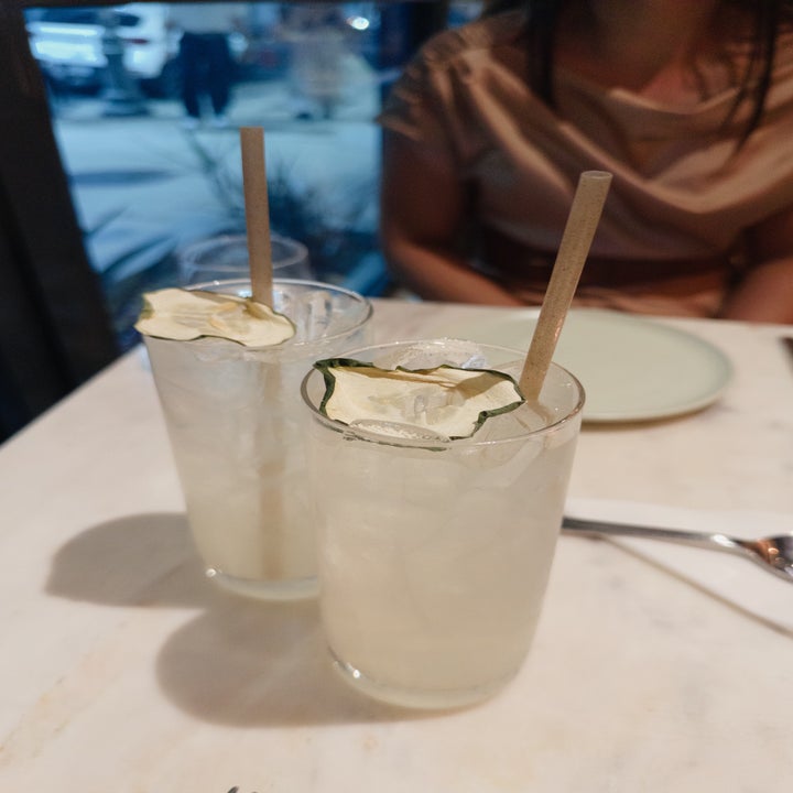 Two glasses of a clear beverage with ice and garnished with dehydrated lime slices on a table. A person in a casual top sits in the background