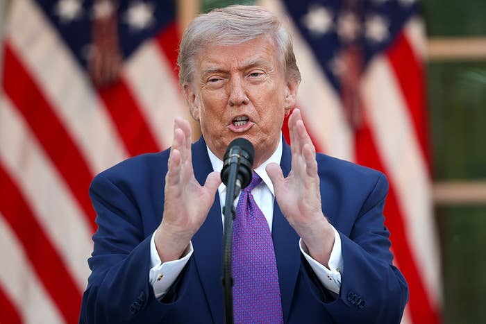 Person speaking at a podium with U.S. flags in the background, wearing a suit and tie, gesturing with hands
