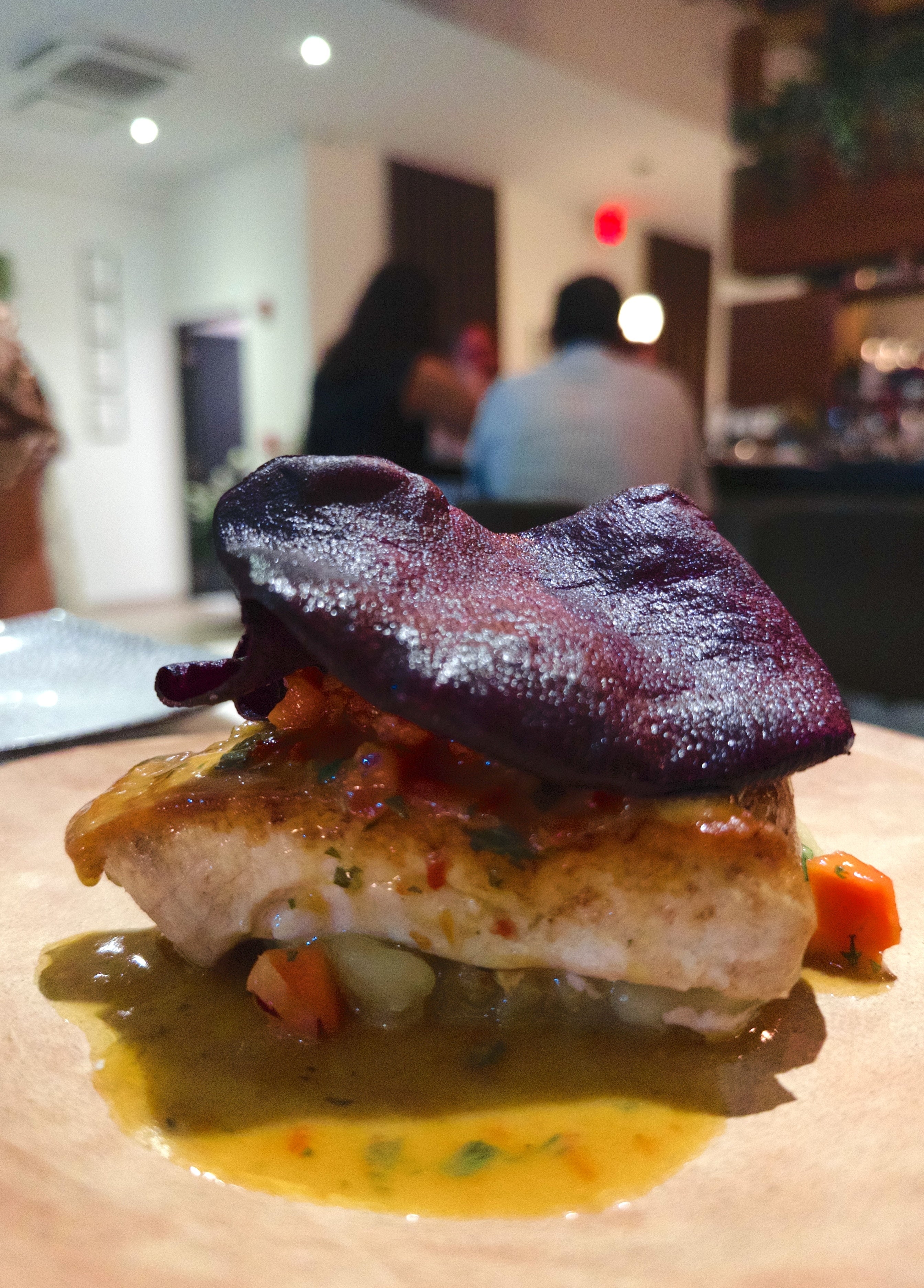 A gourmet dish featuring seared fish topped with vegetables and a crispy garnish, served with a flavorful sauce in a restaurant setting