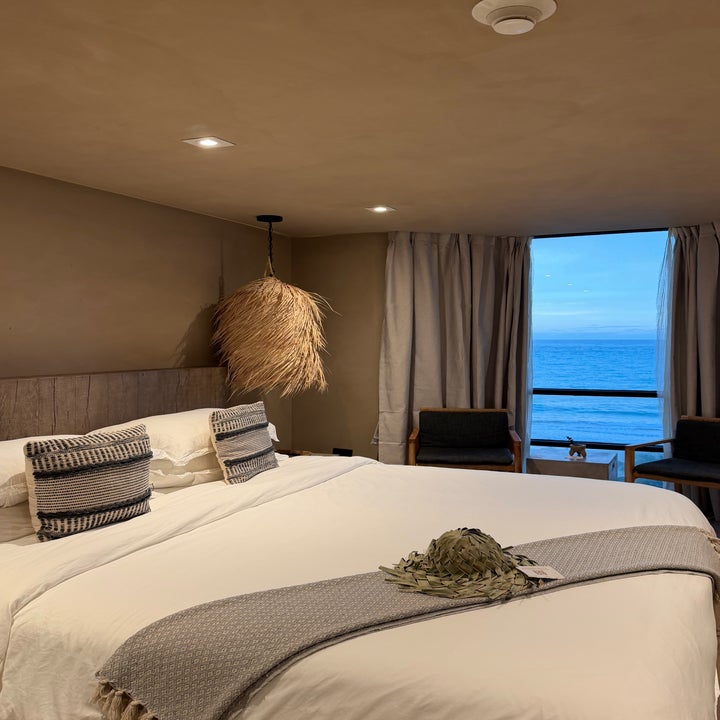 Cozy bedroom with a large bed, woven pillows, a hanging lamp, chairs by a window, and an ocean view