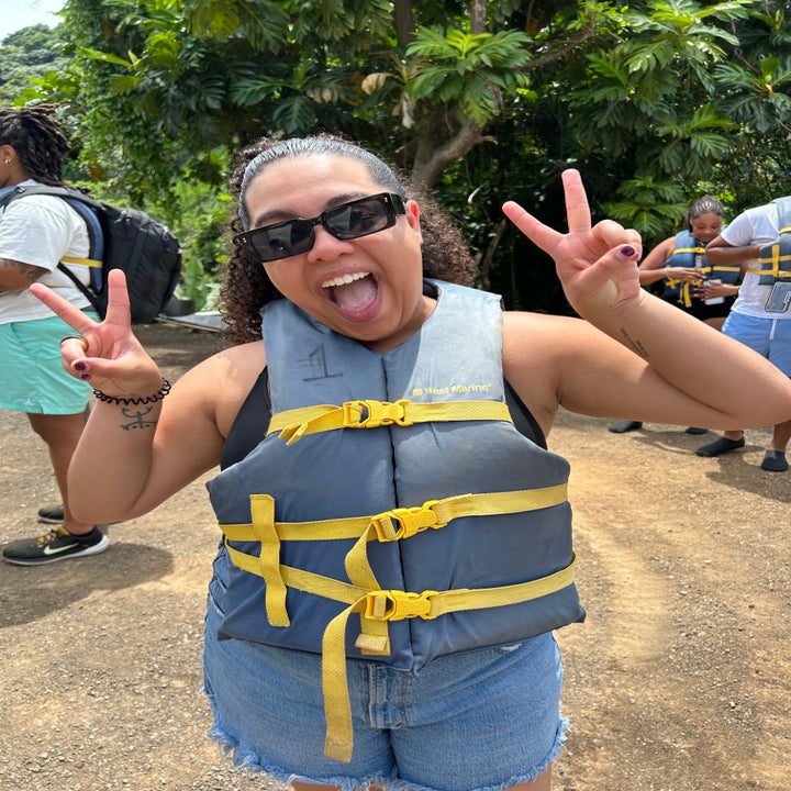 Person smiling and giving peace signs while wearing a life jacket in a sunny outdoor setting. Others in life jackets are in the background