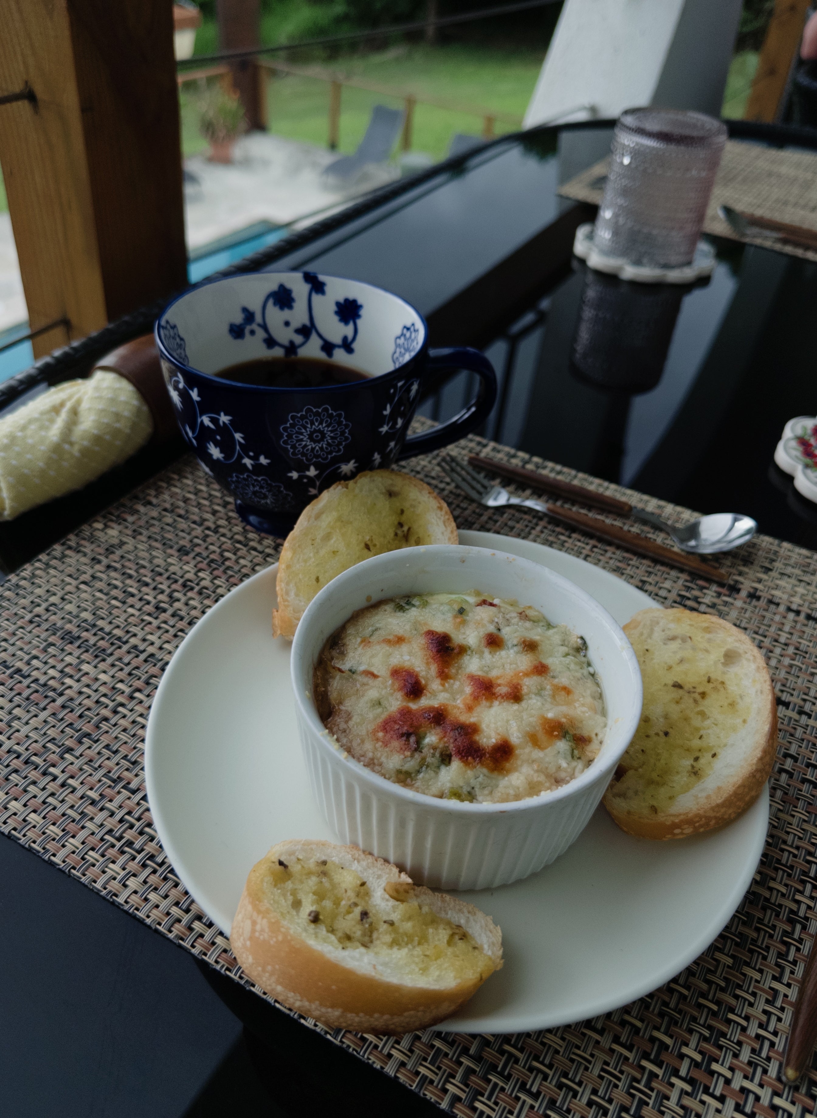 A table setting with a baked dish in a ramekin, surrounded by three slices of bread, and a decorative cup of tea