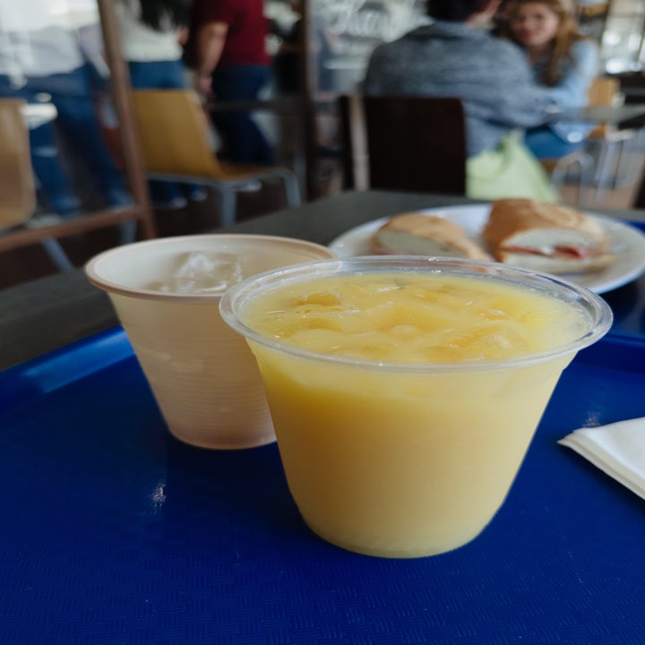 A cup of iced orange juice next to a beverage in a paper cup on a blue tray, with a sandwich in the background