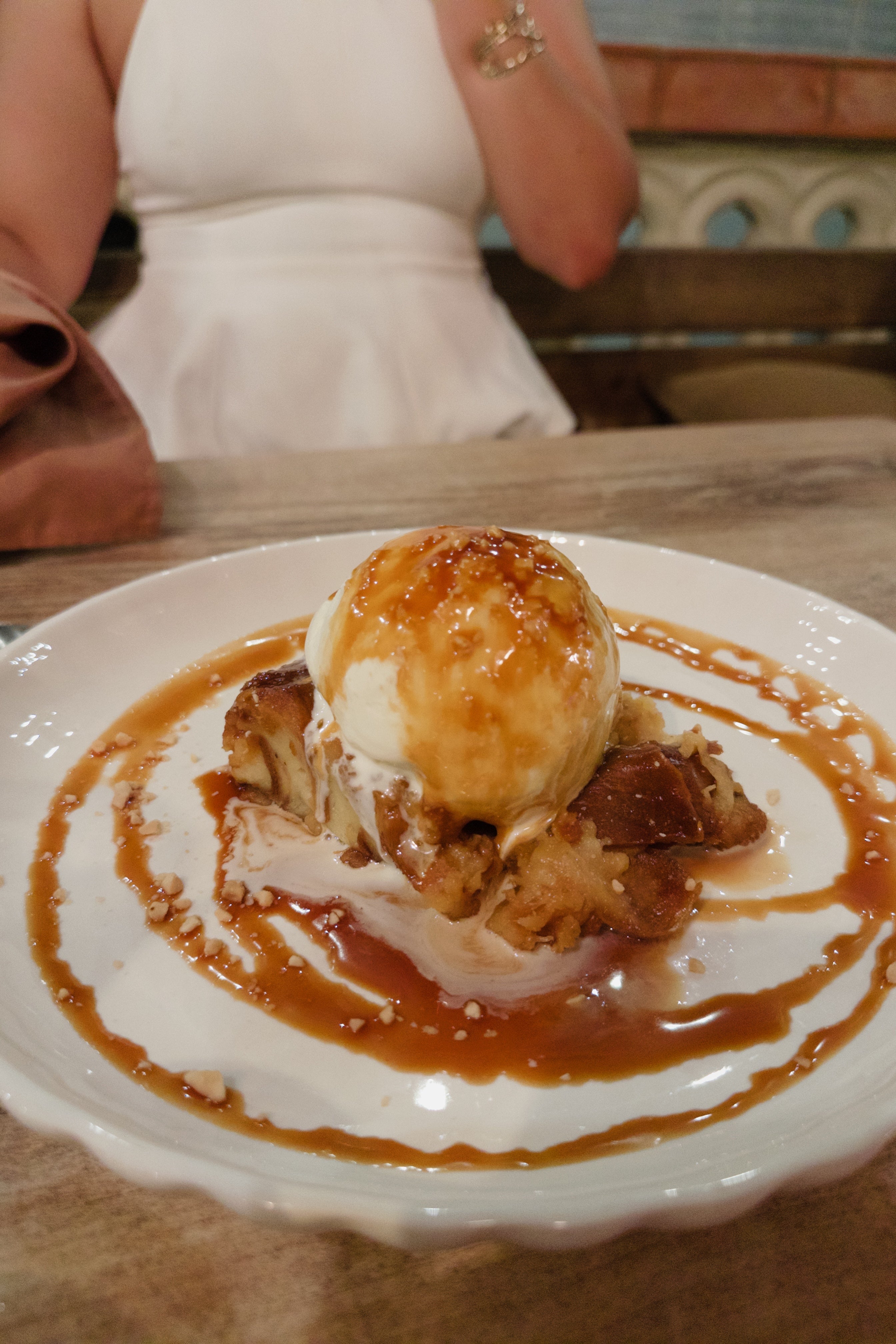 A dessert of bread pudding topped with vanilla ice cream and caramel sauce on a white plate. A person is seated in the background