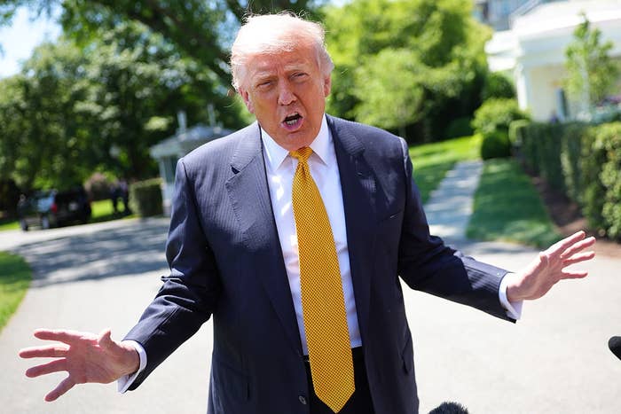 Person in a suit and tie, standing outdoors, speaking to the press with expressive hand gestures