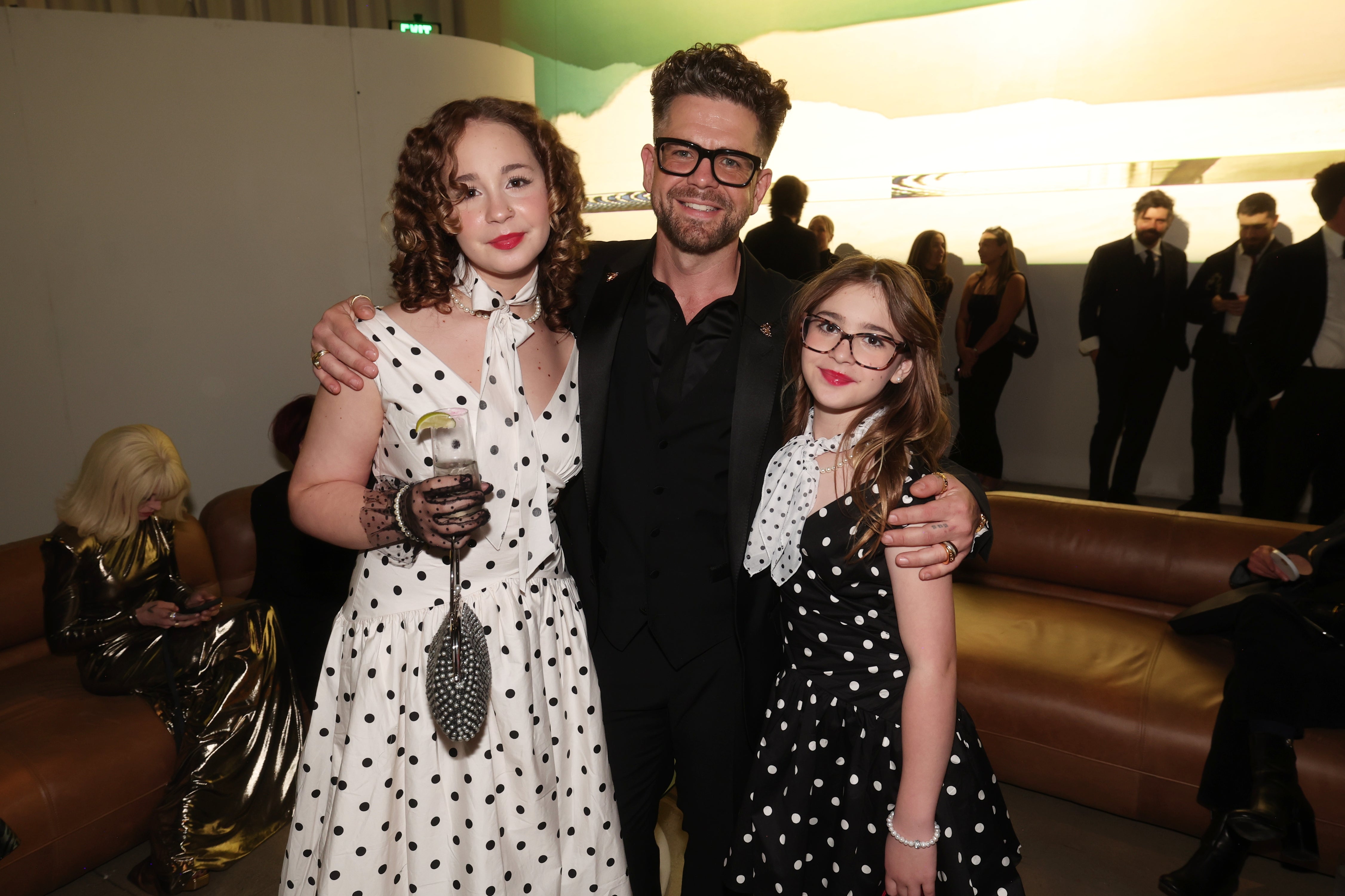 Three people pose at an event, wearing stylish black-tie attire. Two are in polka dot dresses with one person in a suit and glasses