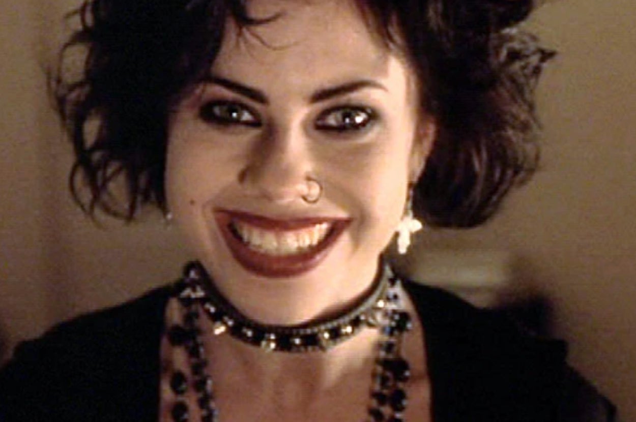 Person smiling widely, wearing dark makeup, a studded choker, cross earrings, and layered necklaces. Hair is styled in a voluminous, edgy manner