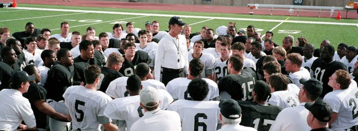 A coach addresses a large group of football players in practice gear on a field, huddled around him attentively