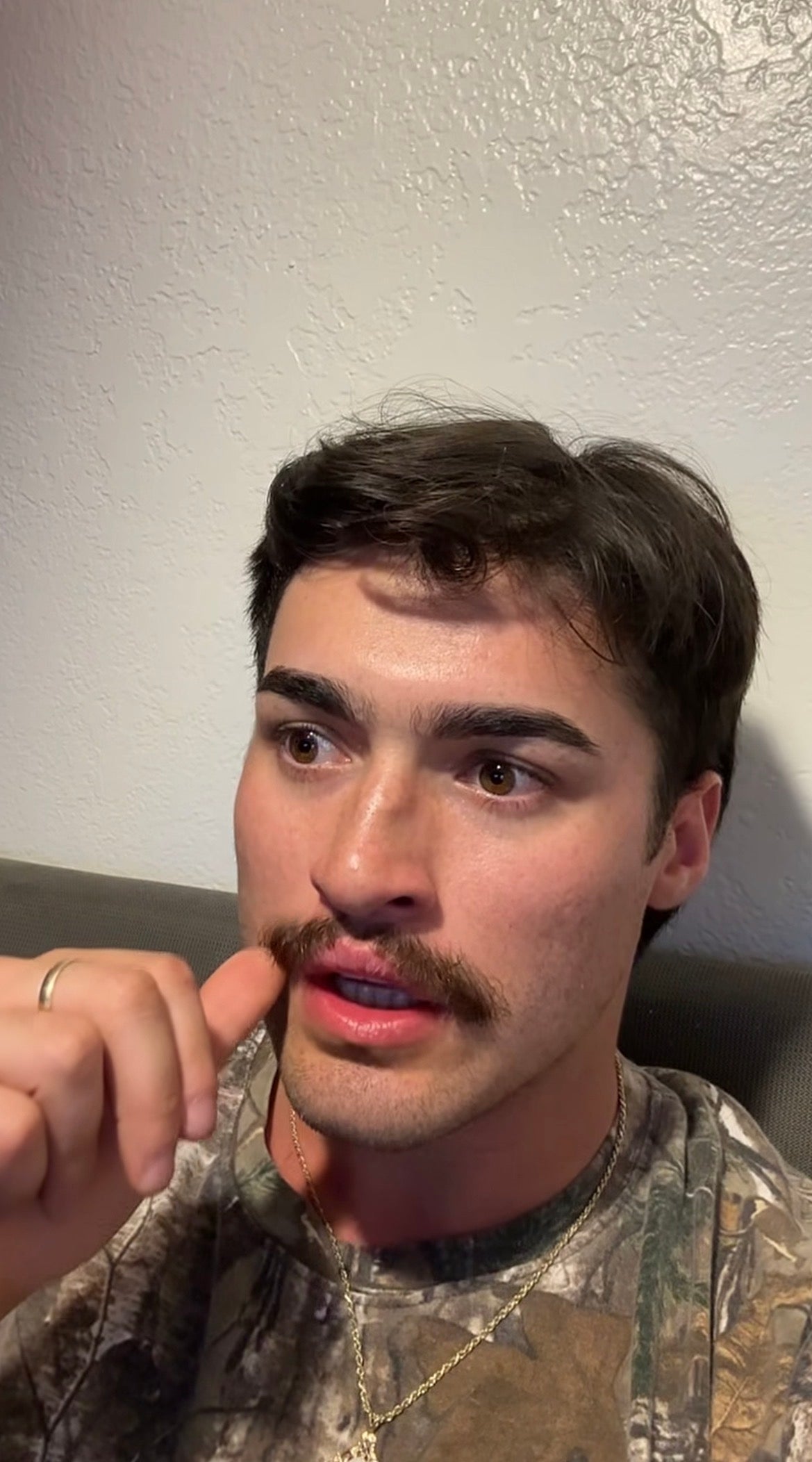 A person with a mustache wearing a camo-patterned shirt, seated indoors, appears to be contemplating something