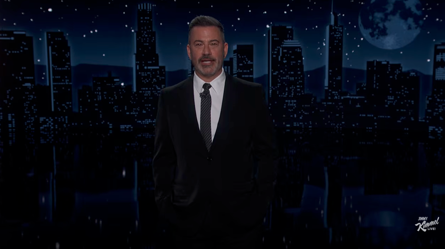 Person in suit on nighttime cityscape set, standing with hands in pockets, speaking or hosting
