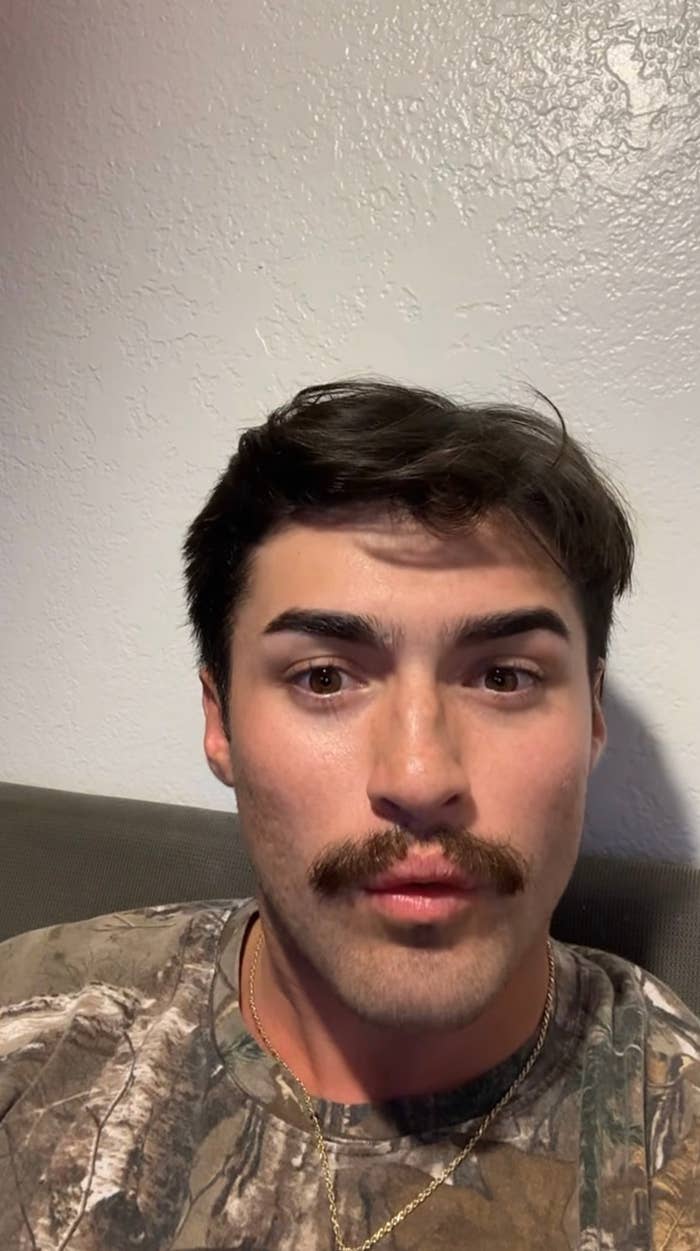 A person with a mustache takes a selfie. They are wearing a camouflage-patterned top and a necklace, sitting against a plain wall